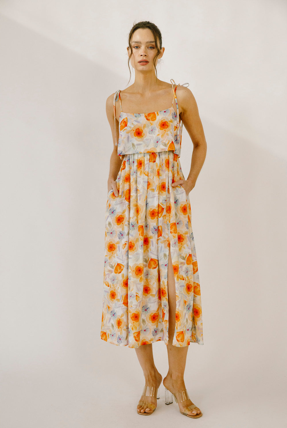 Model wearing a colorful floral print midi dress with a soft watercolor effect, self-tie straps, side pockets and a leg slit against a neutral wall.