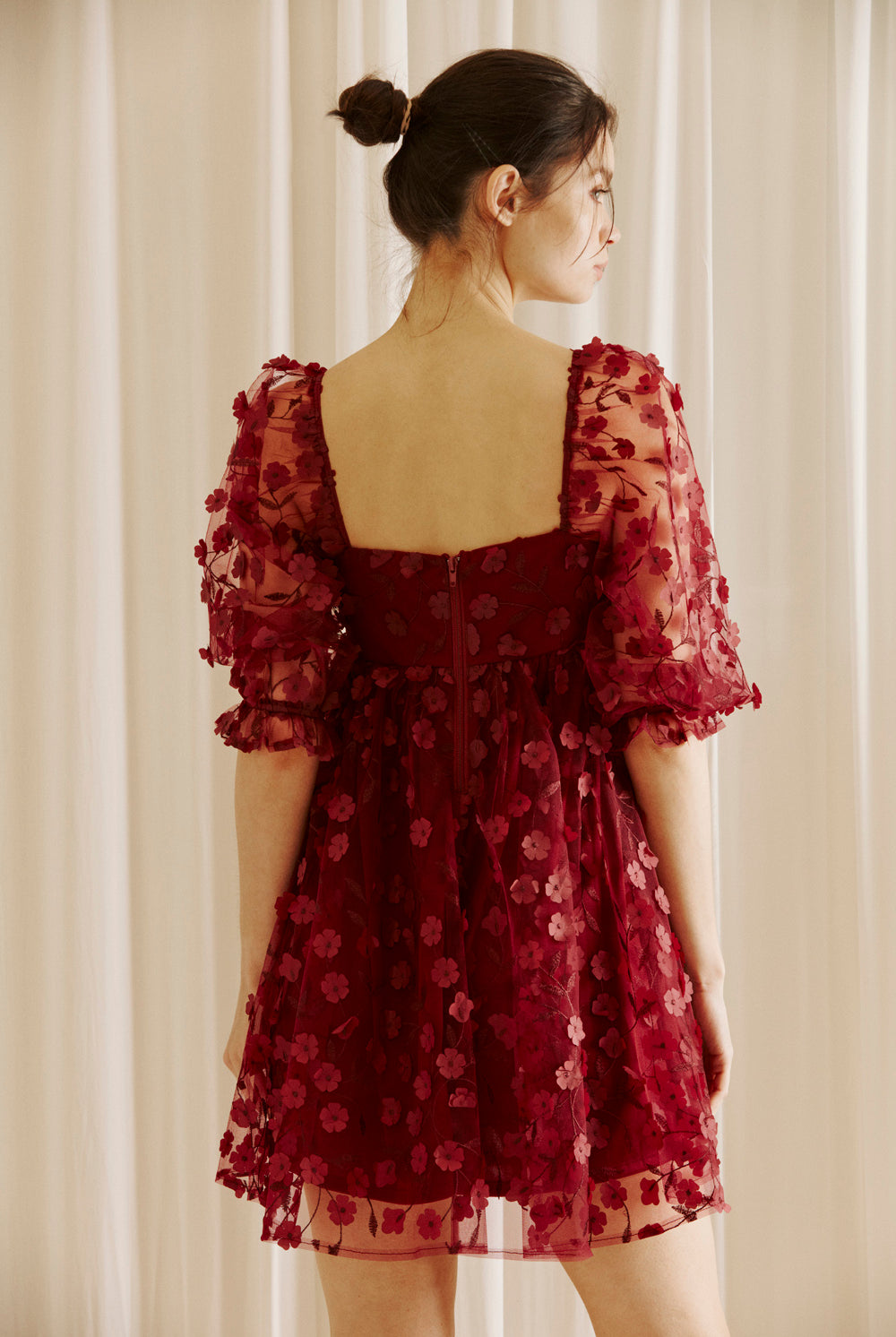 Back view of a woman wearing a burgundy dress with 3D floral detail throughout against a white curtain background