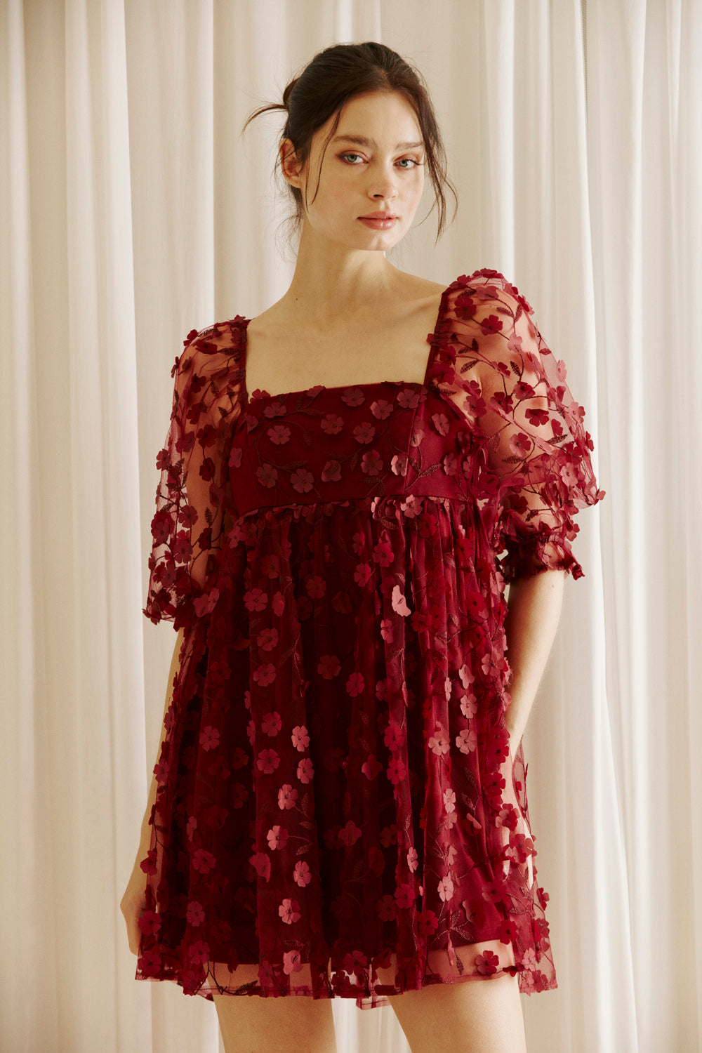 Woman wearing a burgundy dress with 3D floral detail throughout against a white curtain background