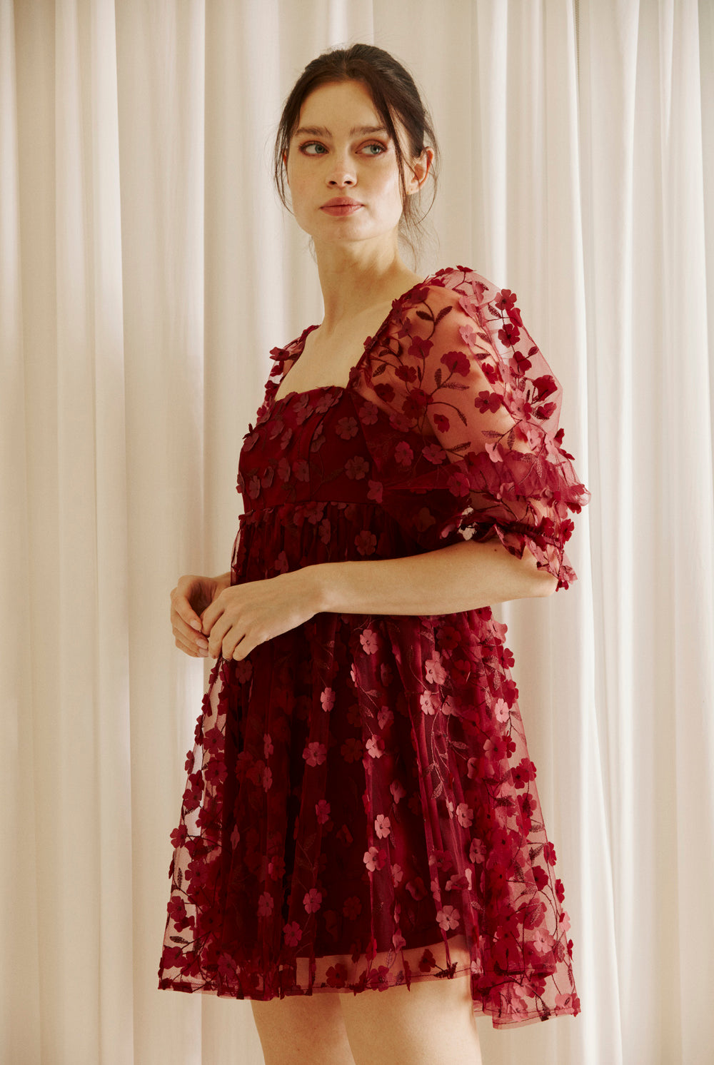 Side view of a woman wearing a burgundy dress with 3D floral detail throughout against a white curtain background