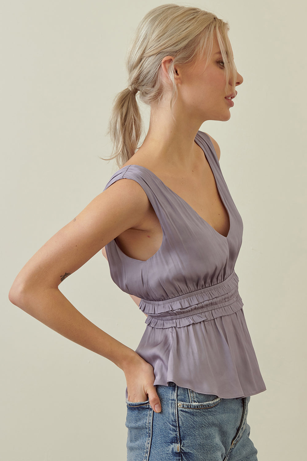 A woman wearing a lavender sleeveless peplum top with a gathered waist, paired with blue jeans, shown from a side profile against a plain beige background.