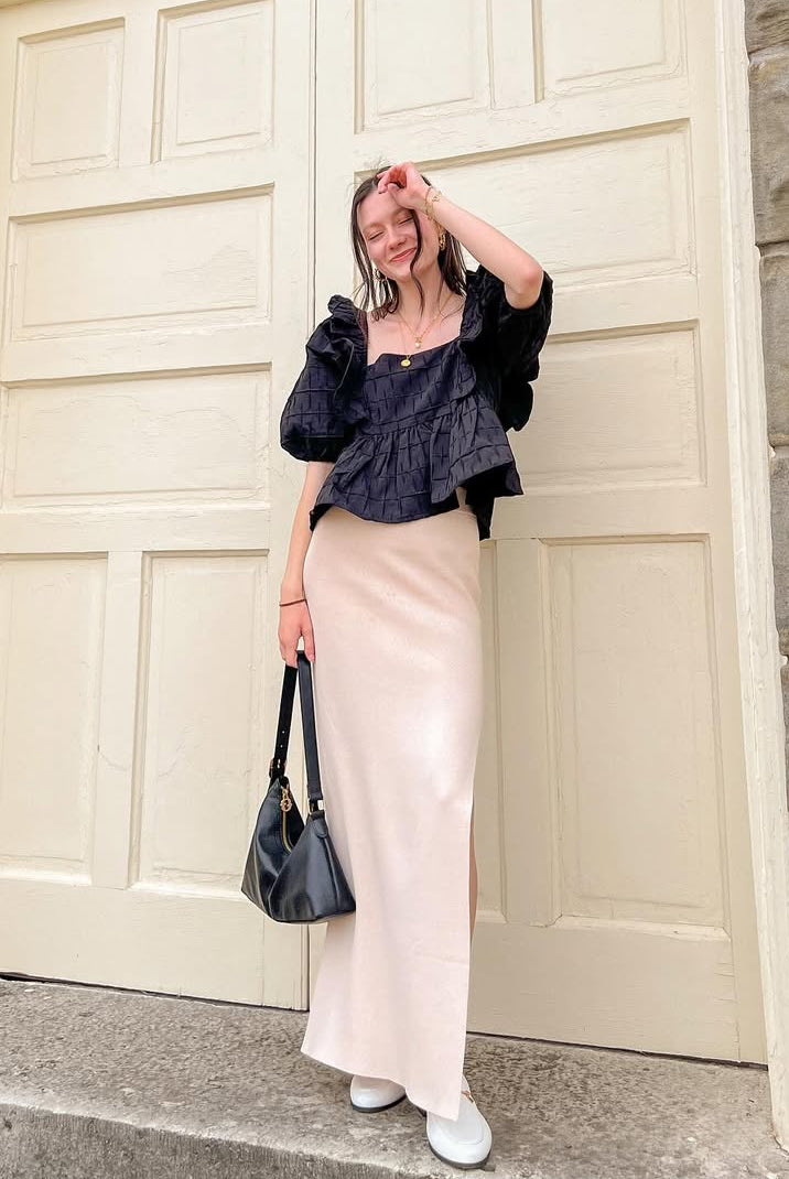 Woman wearing a black blouse and cream skirt posing in front of a large door with stone walls.