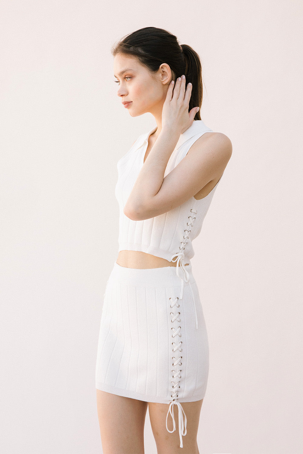 Side view of a woman wearing a white sleeveless top and skirt set with lace-up on the side on a light background