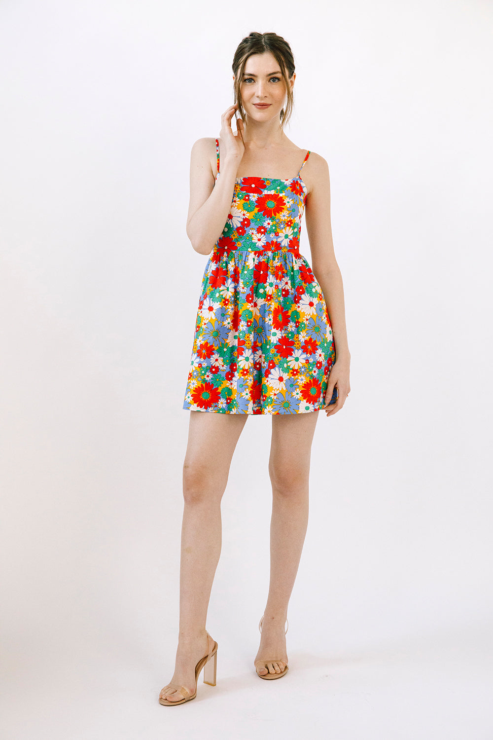 A woman modeling a short floral print mini dress with thin straps and a gathered waist, paired with beige high-heeled sandals, against a plain white studio background.