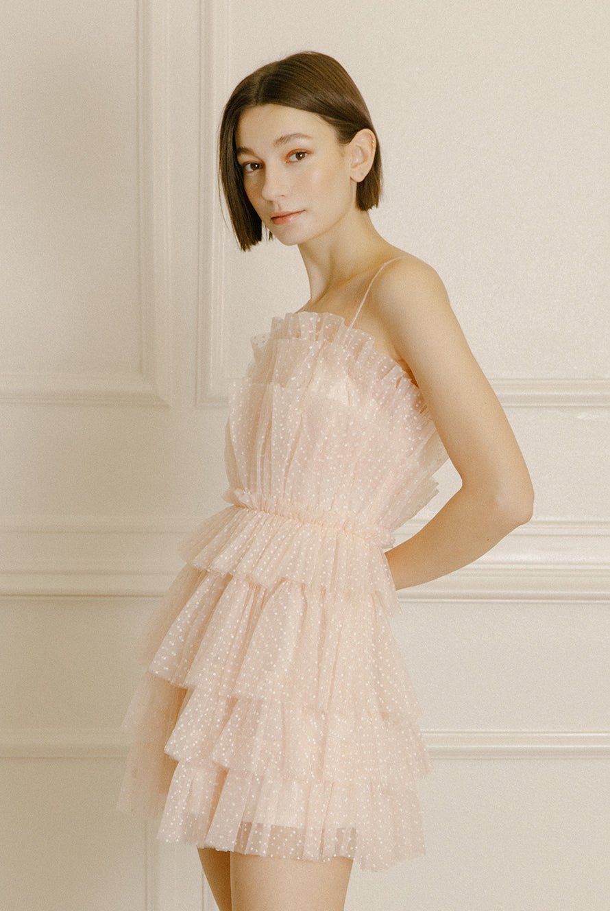 Side of a woman wearing a soft pink mini dress with white dots and tiered tulle on a beige background