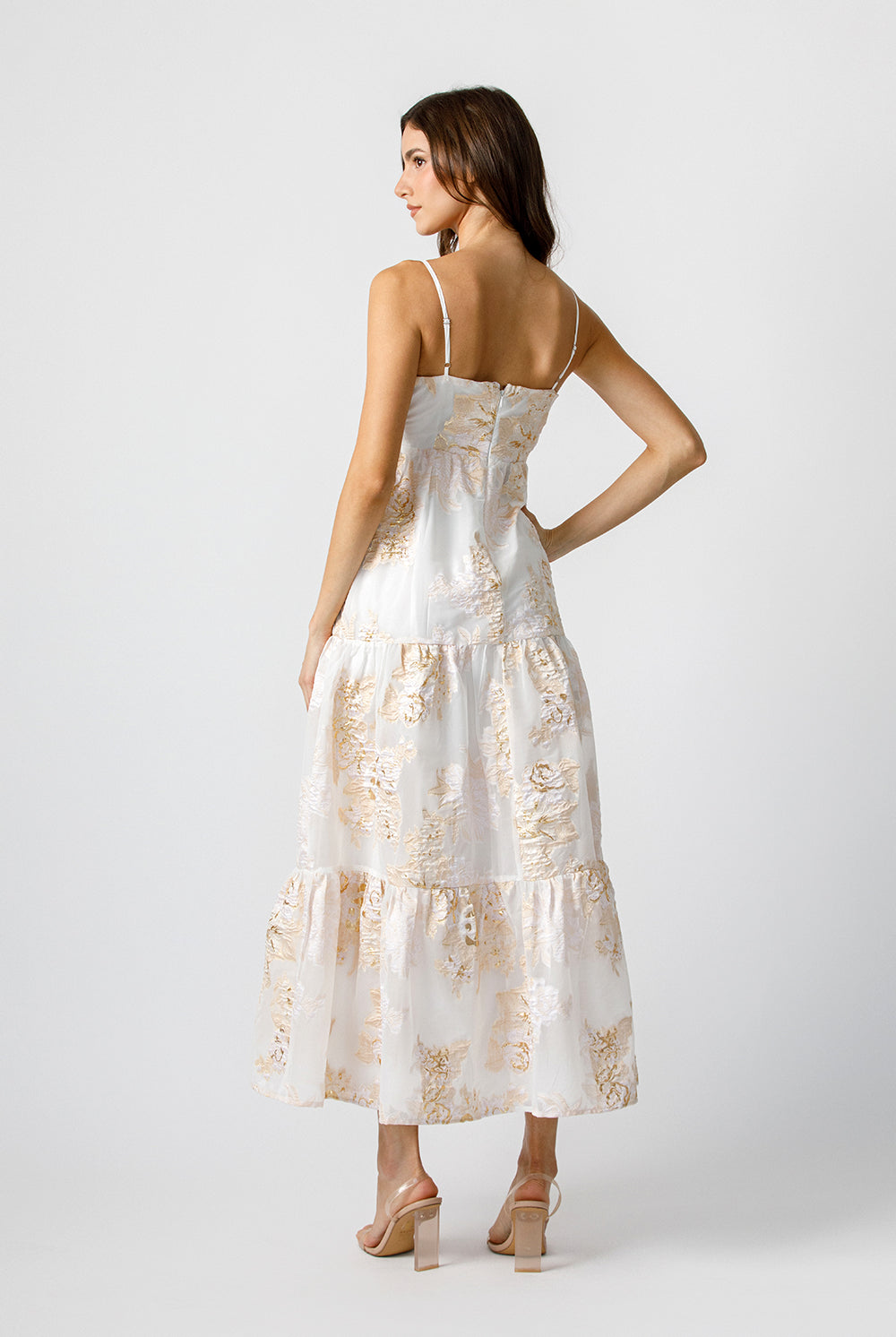 Back view of a woman wearing a white dress with gold floral pattern on a white background