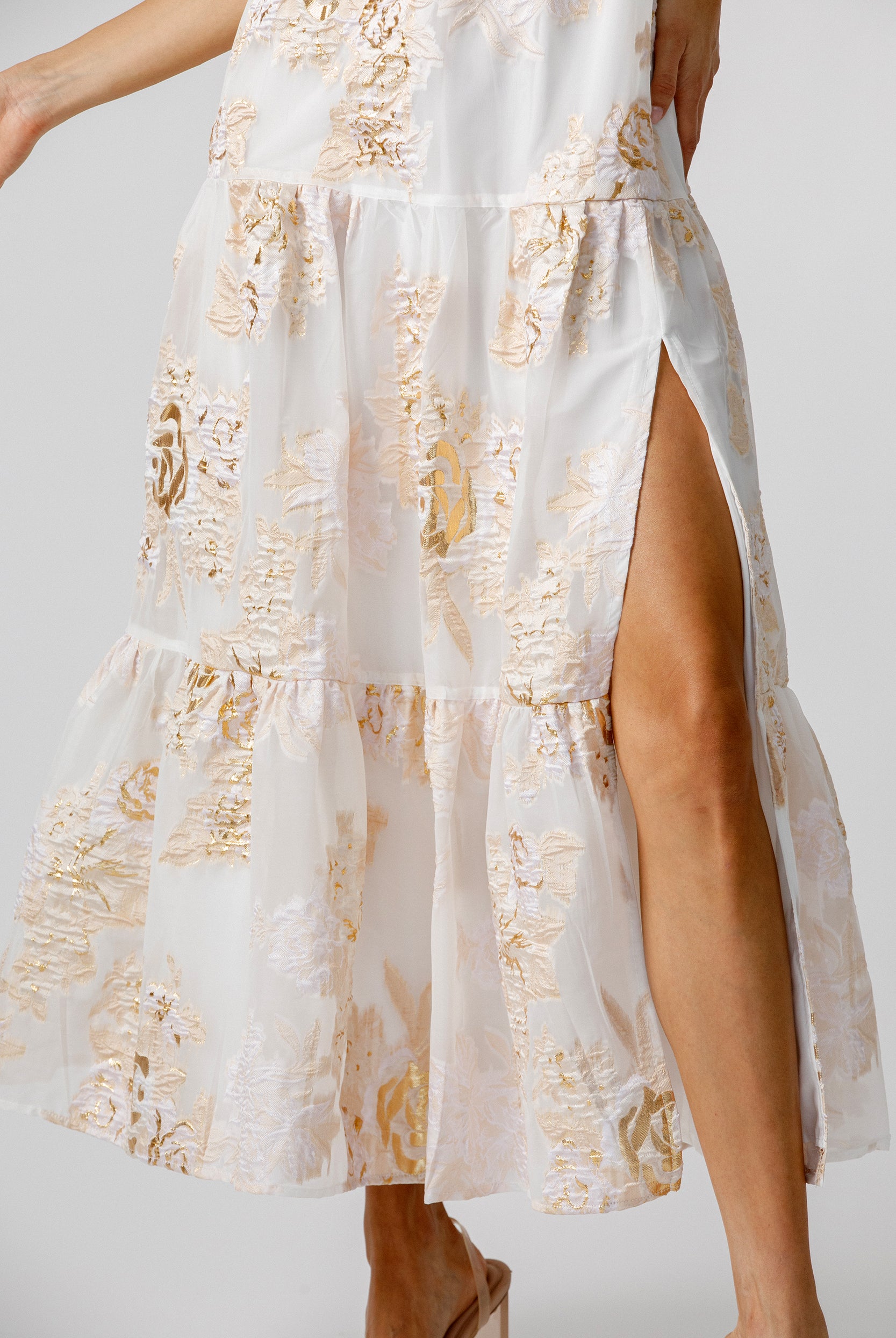 Details of a white dress with gold floral pattern and a slit on a plain background
