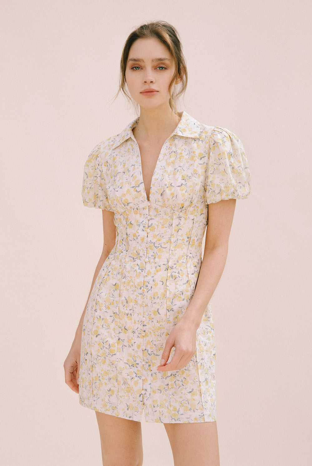 Model wearing a cream dress with a floral pattern, center buttons, a bodycon fitted body and vertical darts from the empire waist down the dress (creating a corset-like detail).