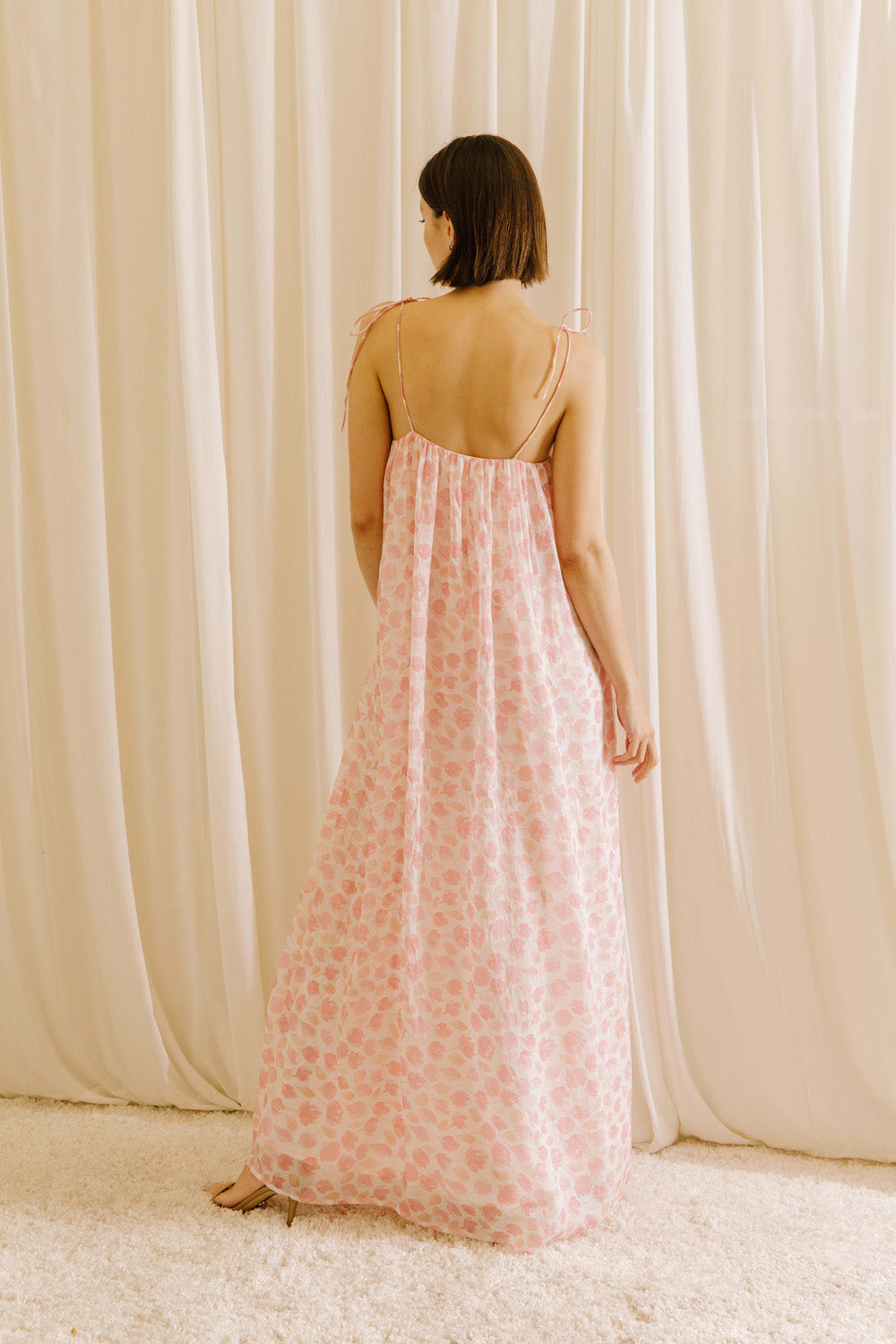 Back view of a model wearing a pink tulip floral dress standing against a beige curtain.