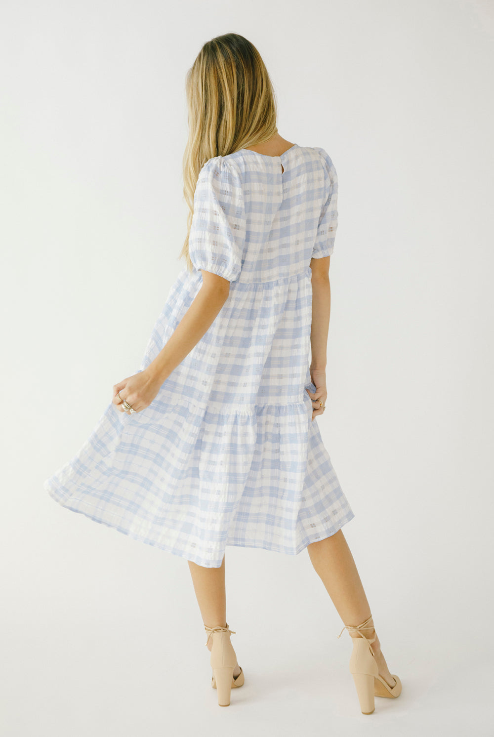 Back view of a woman in a blue checkered dress and beige high-heels against a white wall