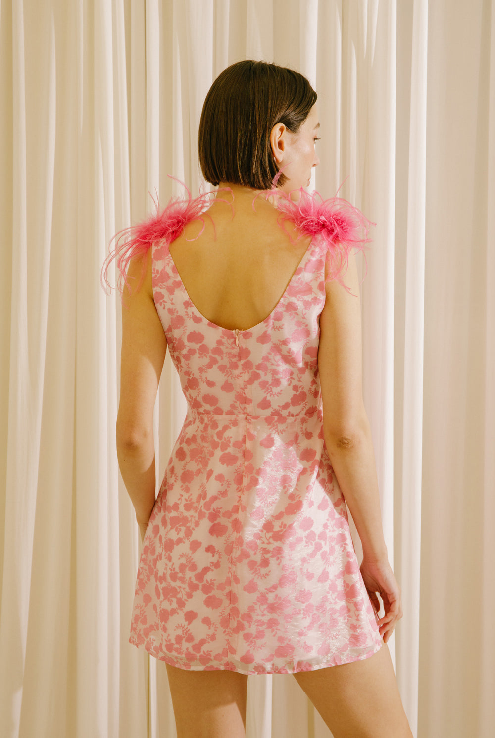 Back view of a woman wearing a white dress with a fun pink pattern and straps adorned with pink feathers against a beige curtain background
