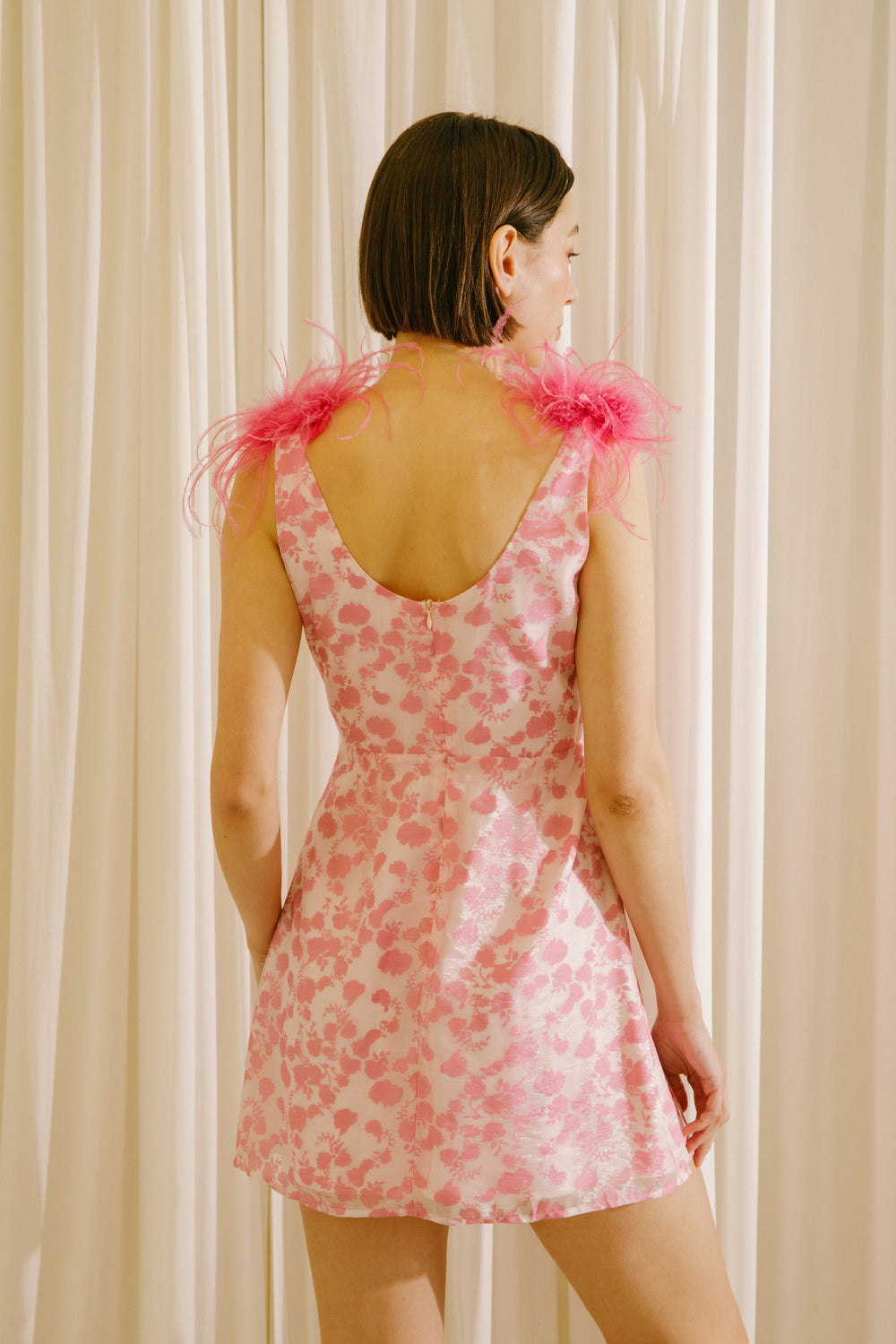 Back view of a woman wearing a white dress with a fun pink pattern and straps adorned with pink feathers against a beige curtain background