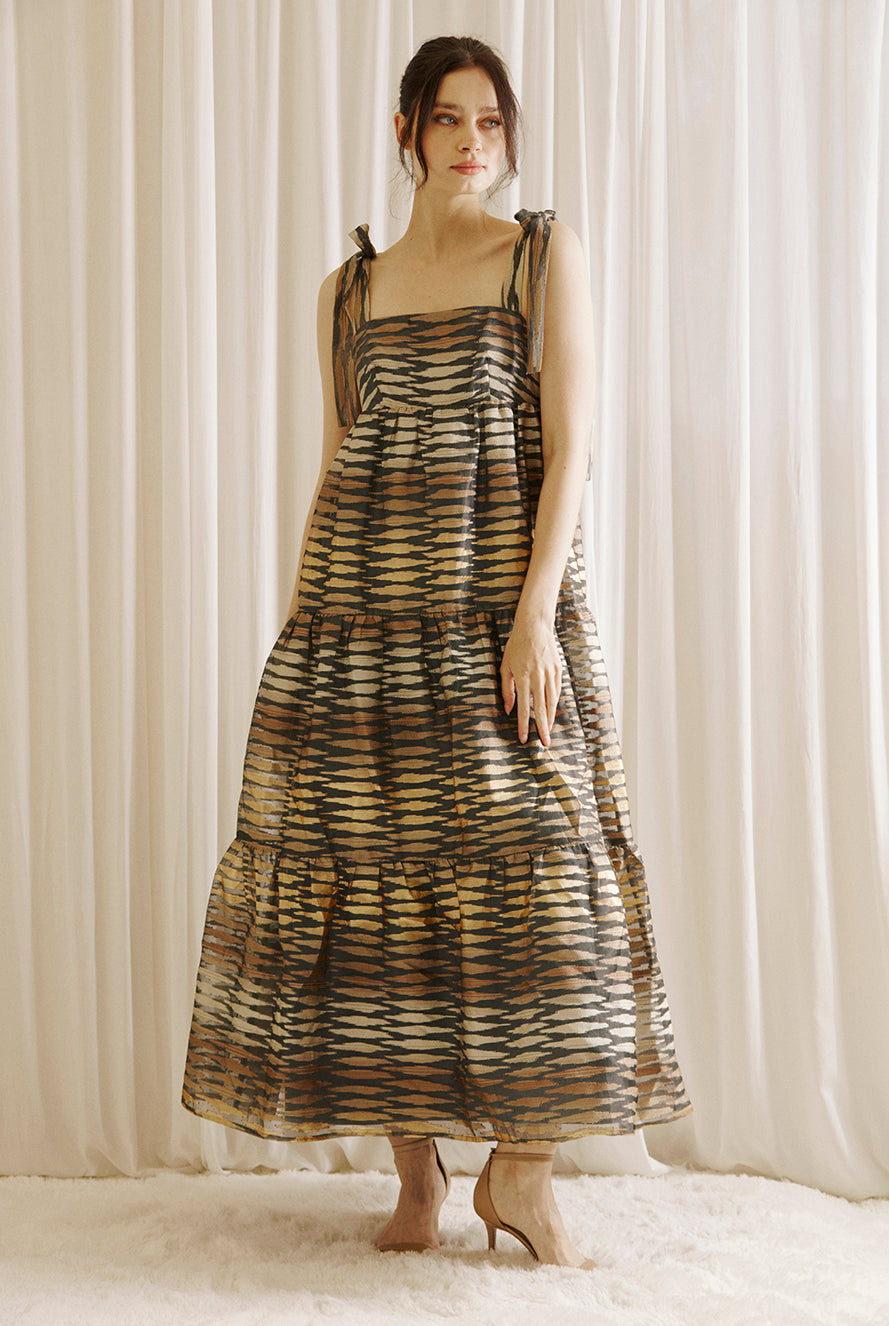 Woman wearing a brown and gold patterned dress standing against a white curtain.