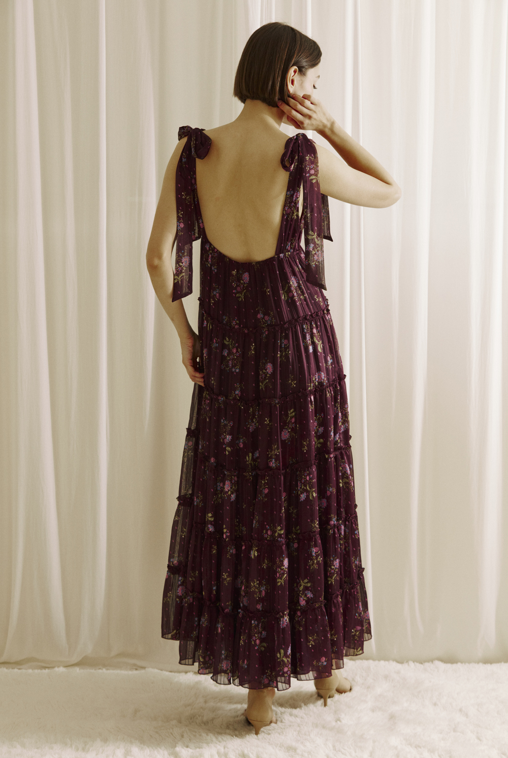 A woman viewed from behind wearing a plum base maxi dress with an allover pink floral print and a low scoop open back on a white background