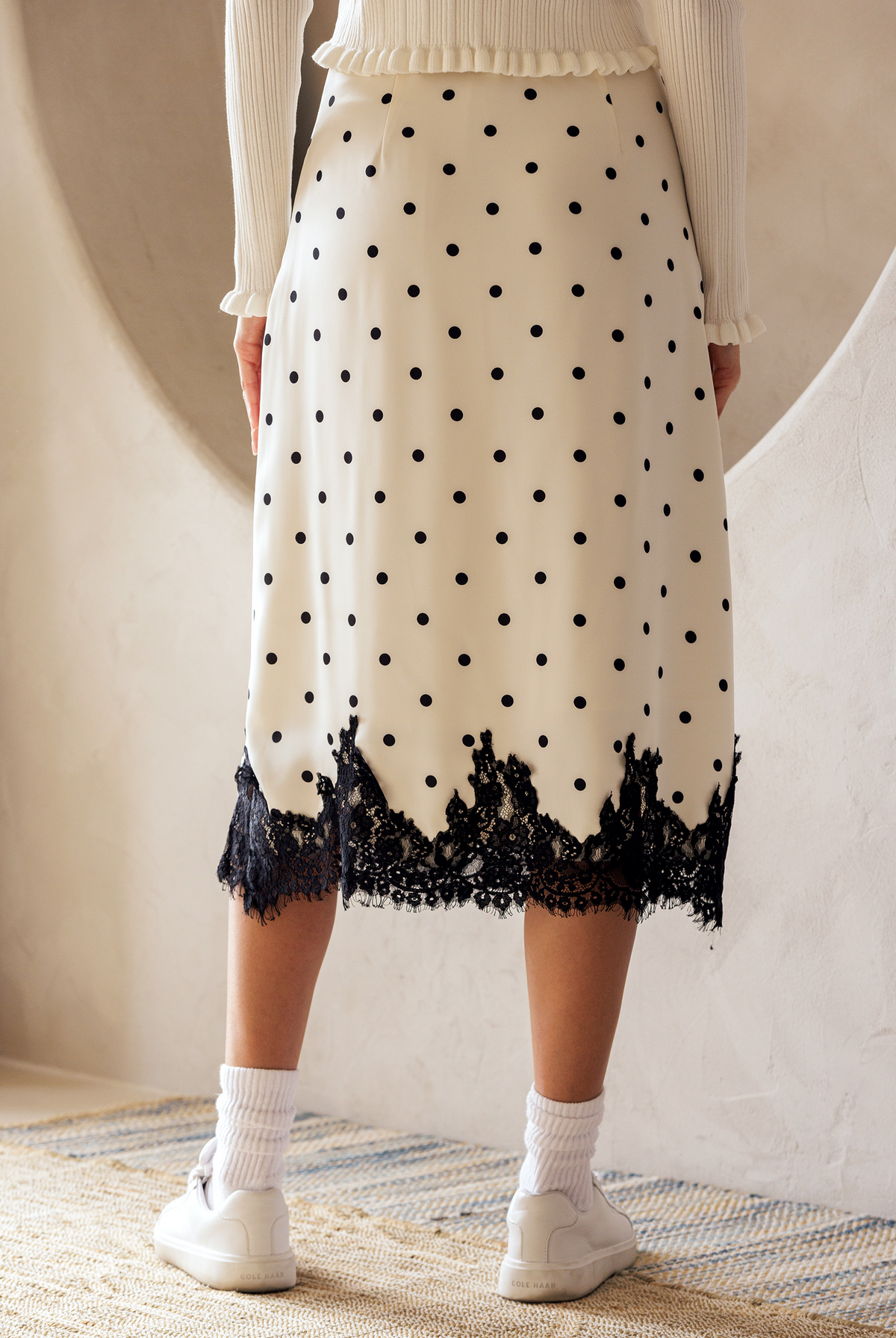 Back view of a person wearing a midi skirt that features an ivory and black polka dot print and a black lace-trimmed hem.
