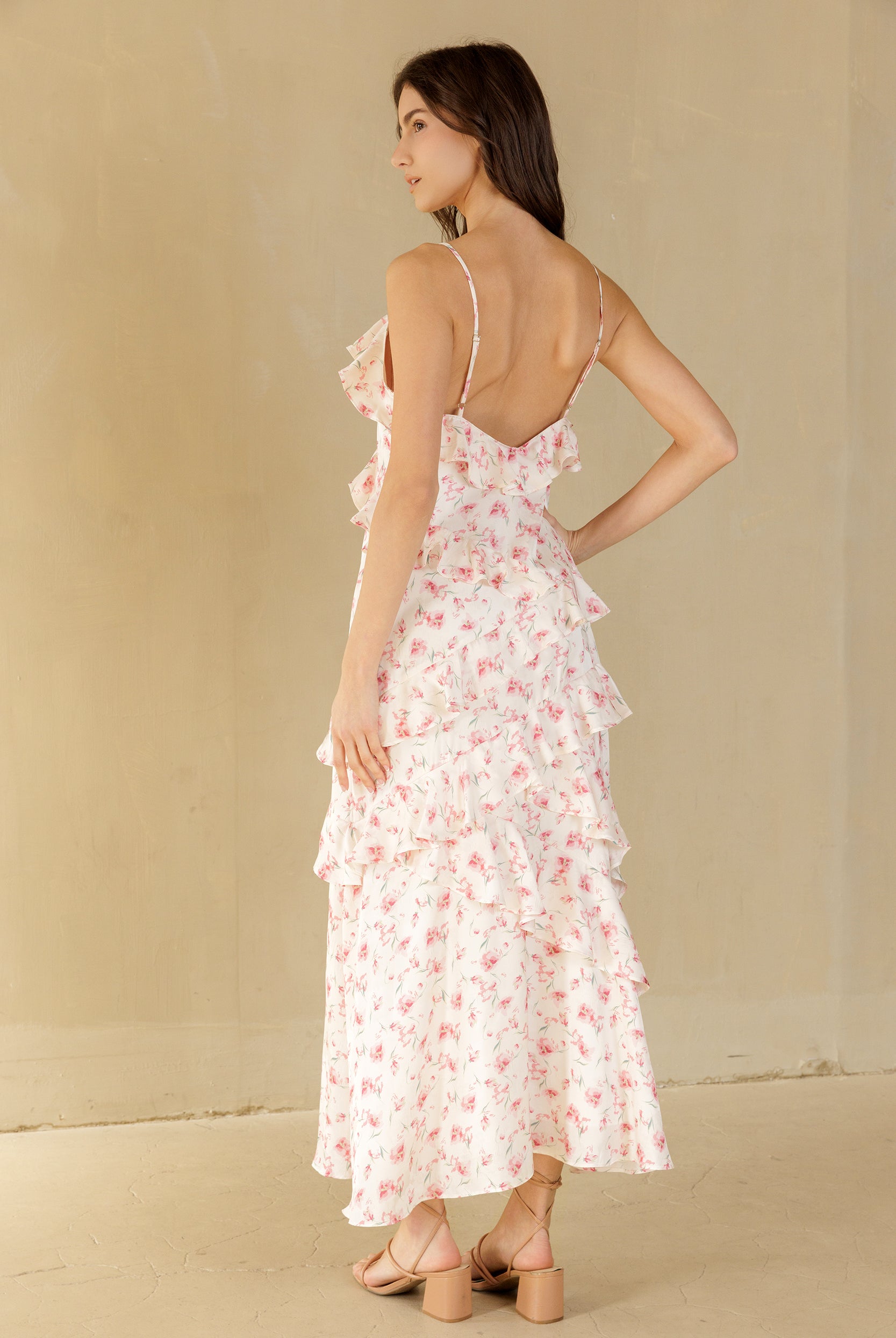 A woman modeling a white tiered maxi dress with a blush floral print, shown from the back against a neutral beige background.