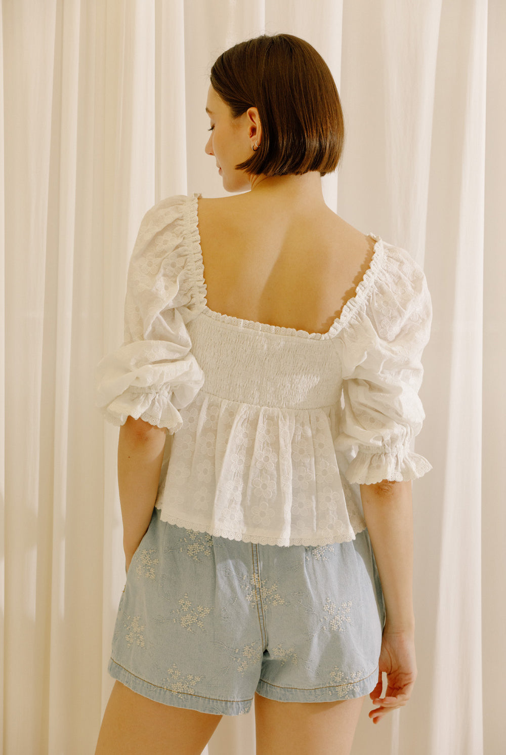 A woman shown from the back wearing a white puff-sleeve lace top with a square neckline and ruffled trim, paired with light blue denim shorts, standing in front of cream-colored curtains.