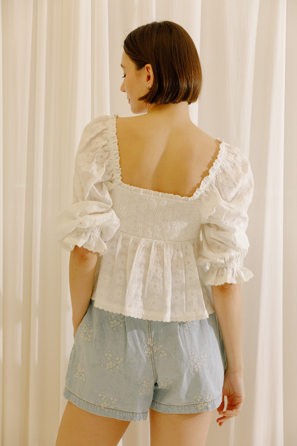 A woman shown from the back wearing a white puff-sleeve lace top with a square neckline and ruffled trim, paired with light blue denim shorts, standing in front of cream-colored curtains.