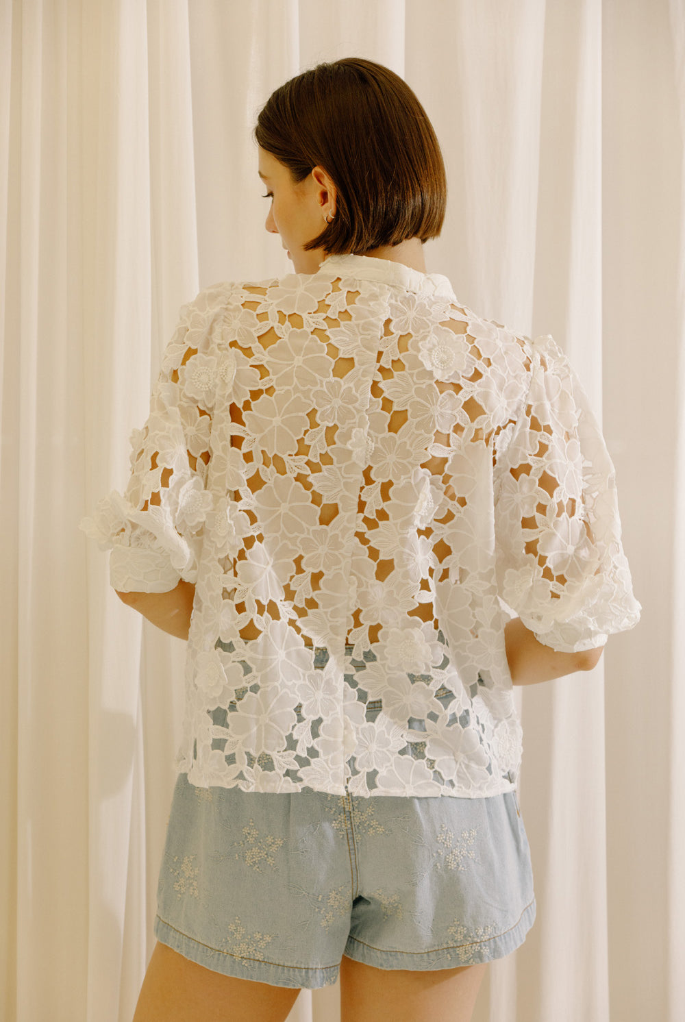 A woman viewed from behind wearing a white eyelet short-sleeve blouse and light blue denim shorts, standing in front of white curtains.