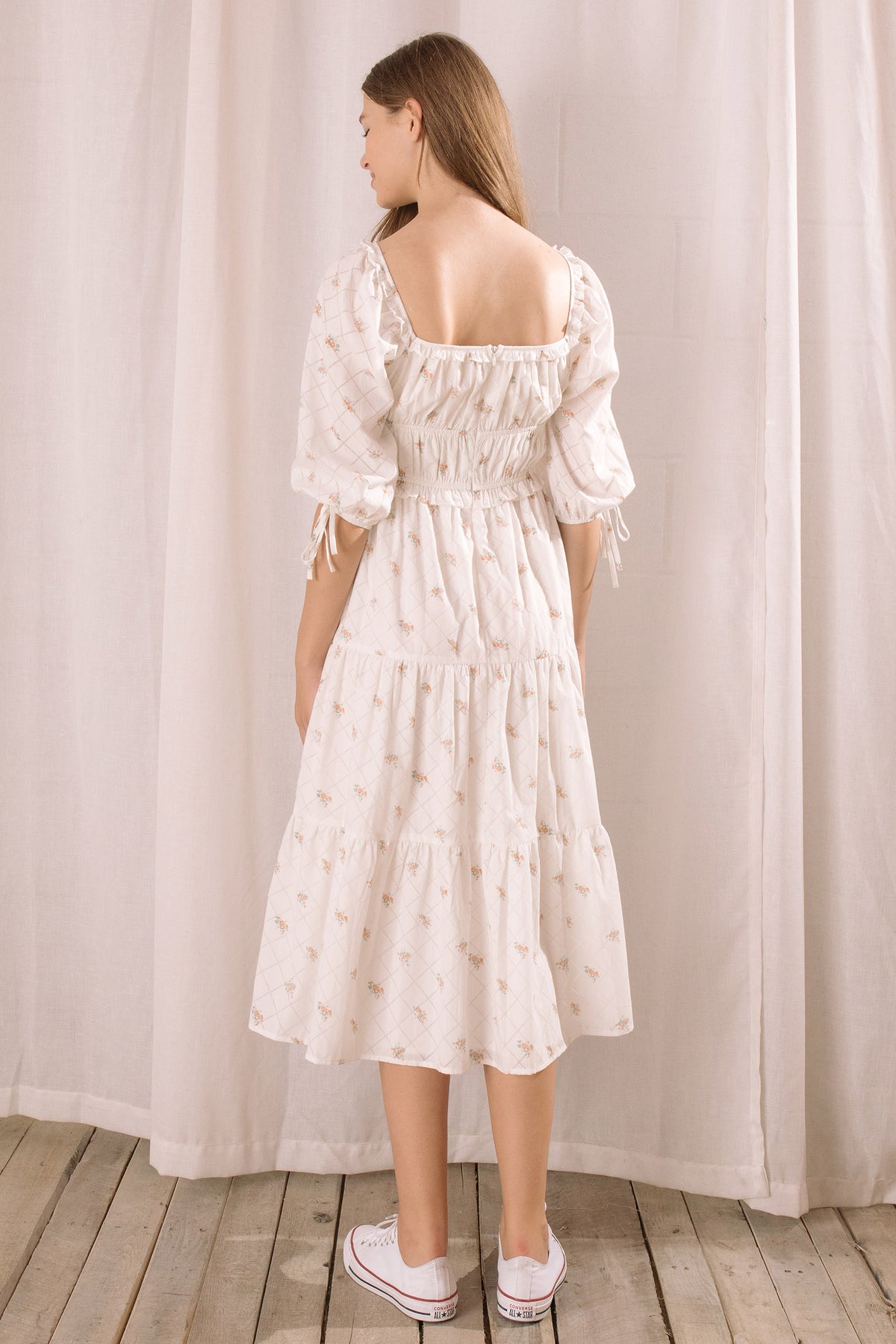 A woman viewed from behind wearing a white dress with puff sleeves against a neutral curtain background