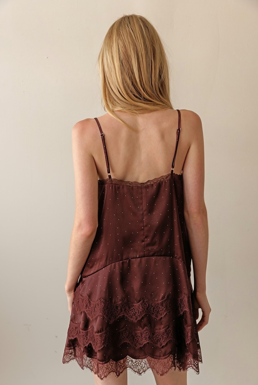 A woman viewed from behind wearing a dark brown spaghetti-strap slip dress with a lace hem, set against a plain beige wall.