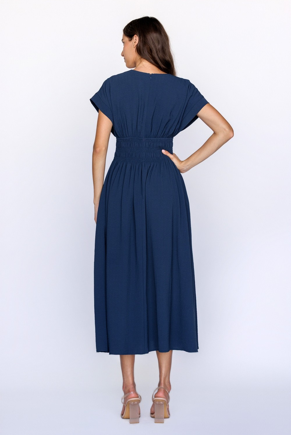 A back view of a model wearing a navy midi dress with a smocked waist and dolman sleeves against a white wall.