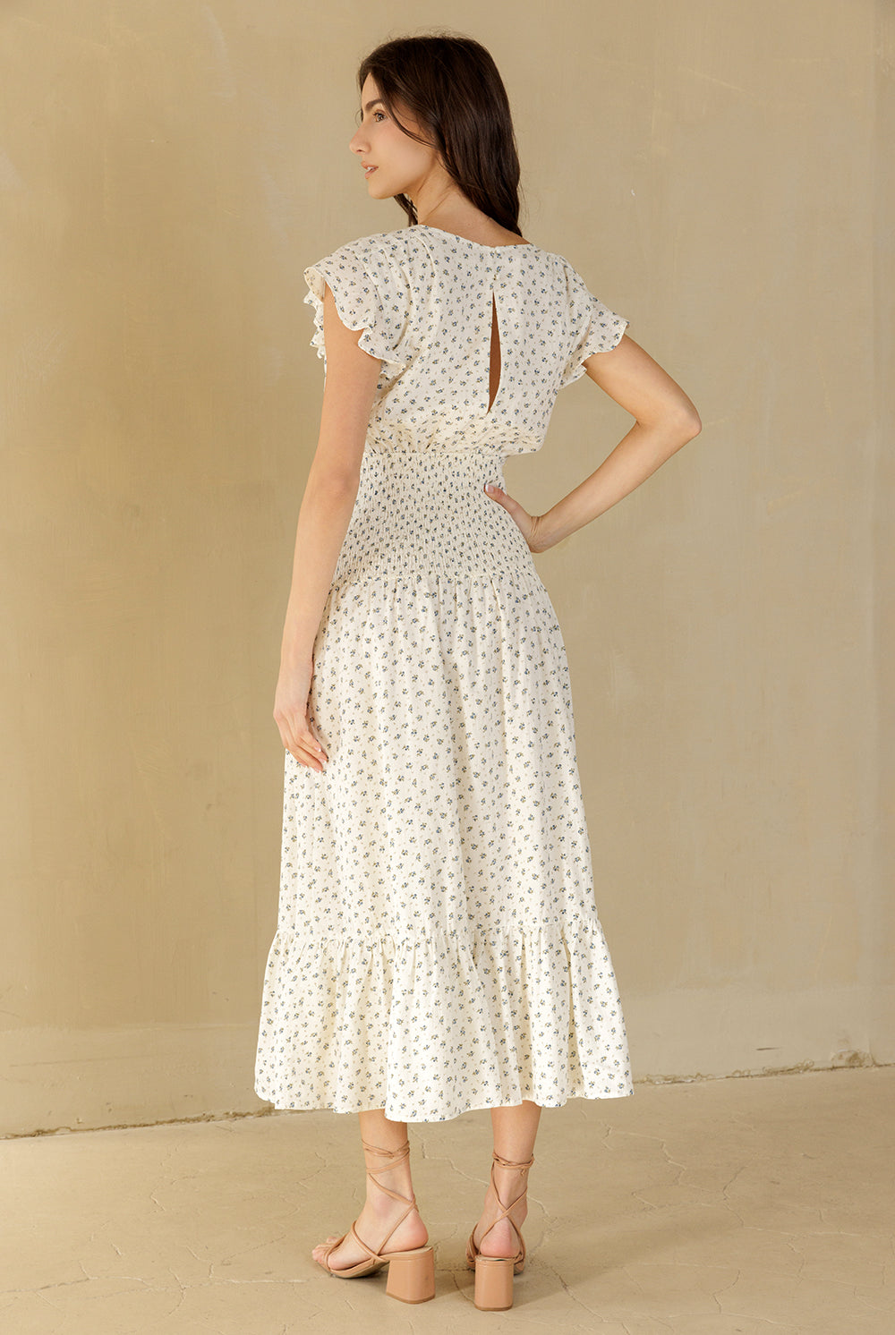 A woman wearing a cream midi dress with a small blue and green floral pattern, shown from the back. The dress features ruffled cap sleeves, a keyhole back with a decorative tie, and a tiered skirt.
