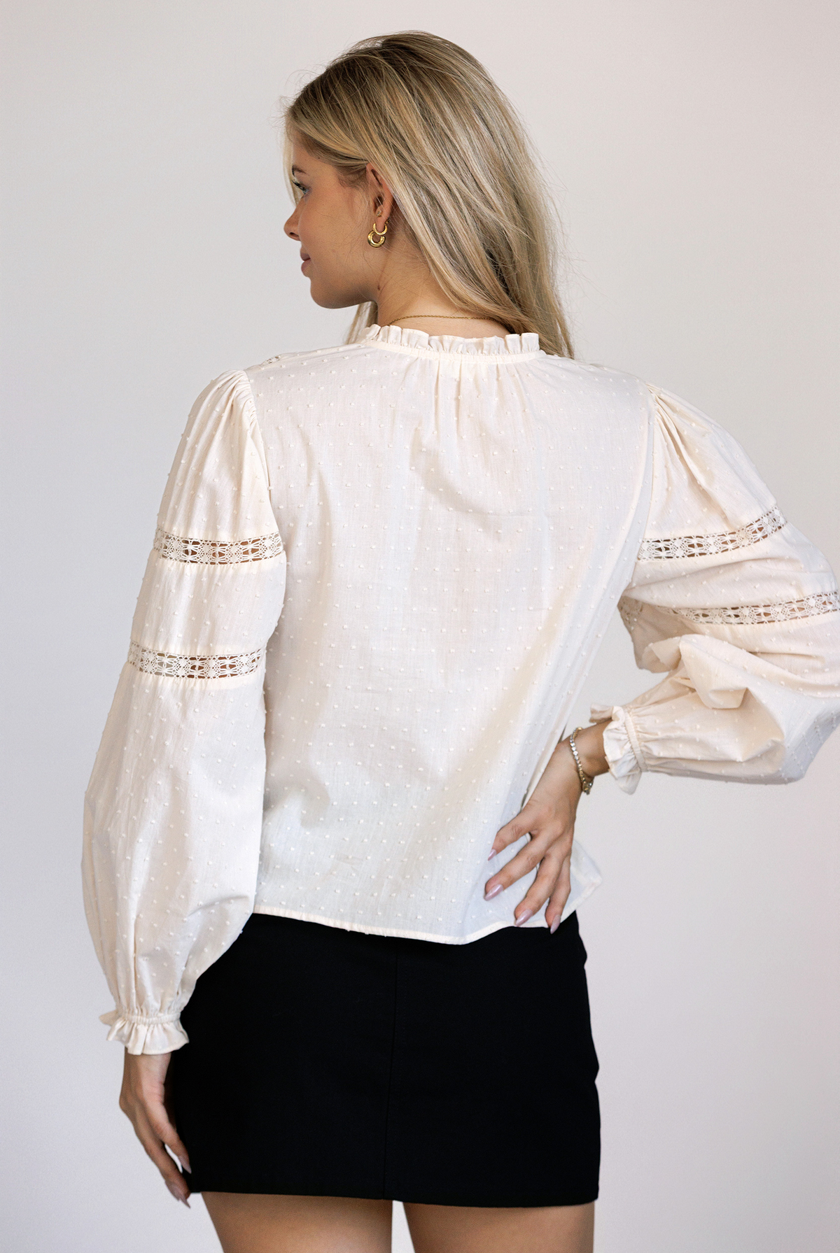 A woman viewed from behind wearing a cream long sleeve top featuring textured Swiss dot cotton, delicate lace paneling, puff sleeves.
