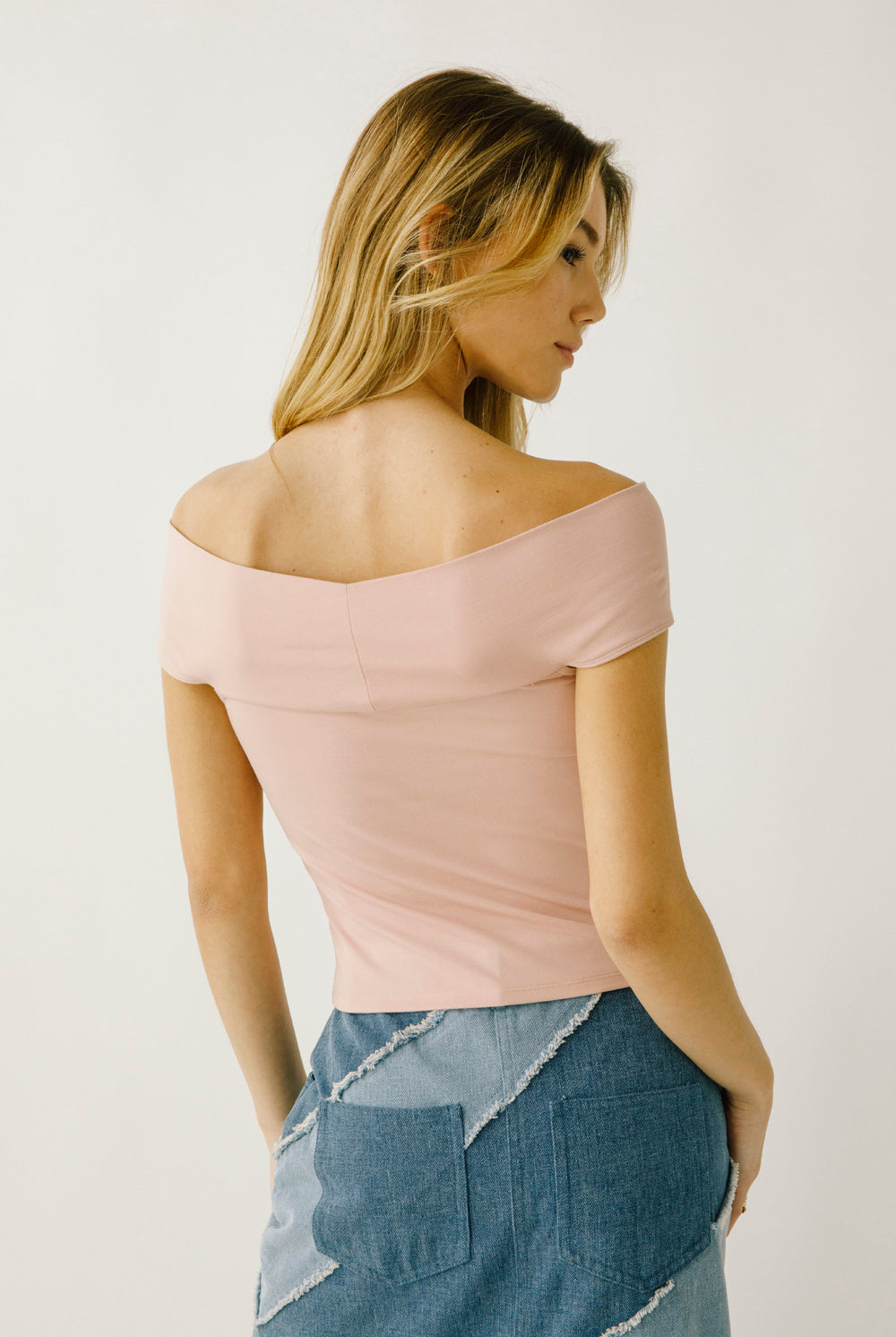 A woman viewed from behind wearing a light pink off-shoulder top and denim skirt on a plain background