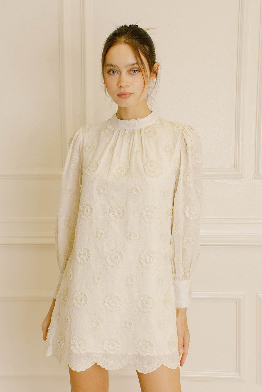 Woman wearing a cream daisy embroidery mini dress against a neutral wall.
