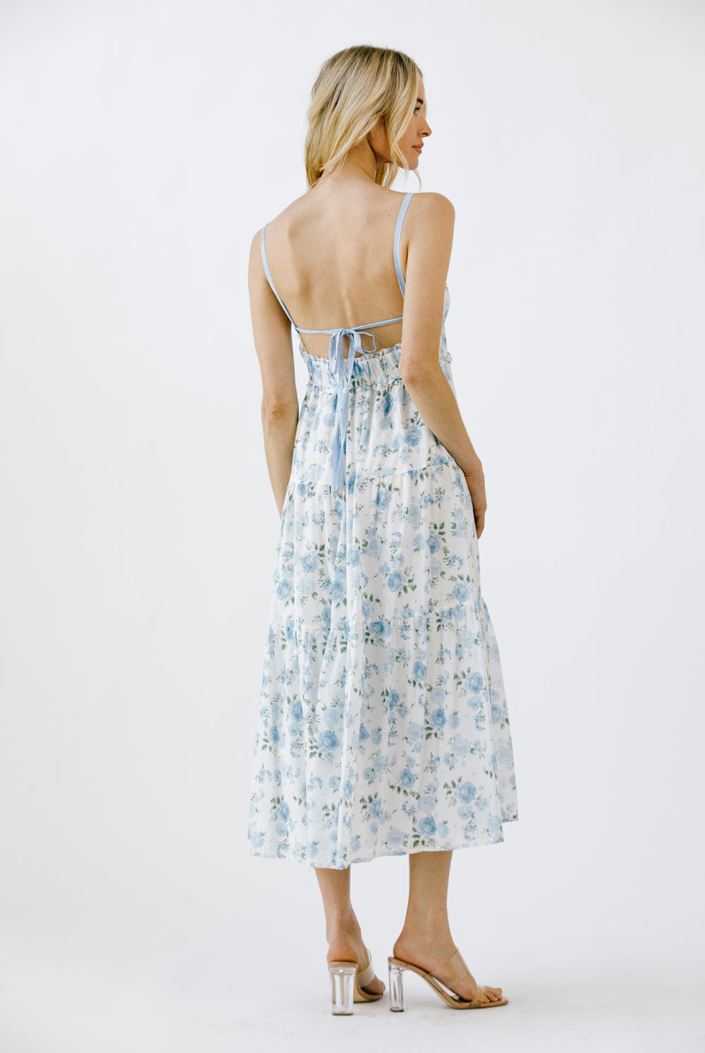 Back view of a woman wearing a blue and white floral midi dress with an open back with self-tie bow on a white background