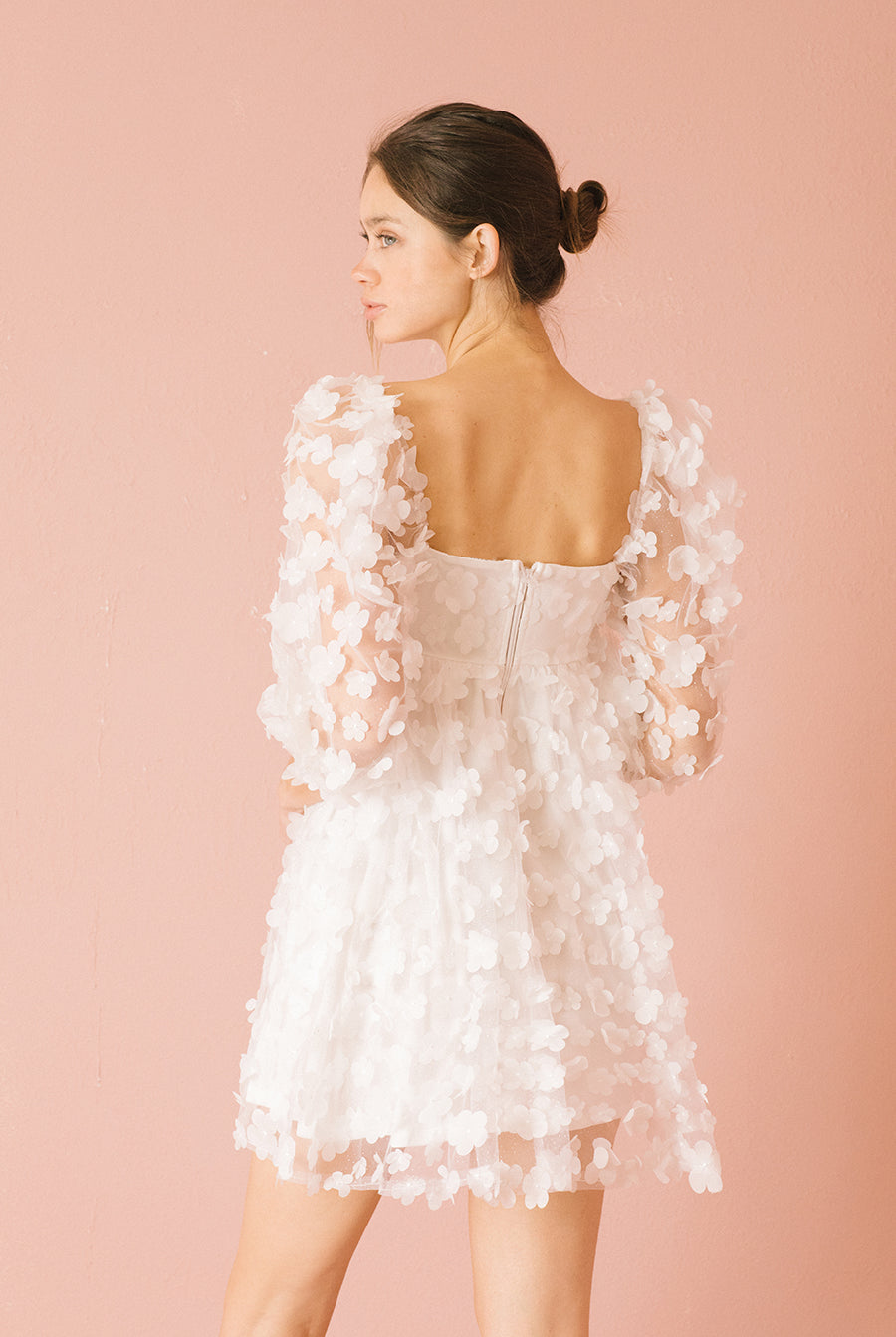 Back view of a woman wearing a white 3D floral mini dress against a pink background