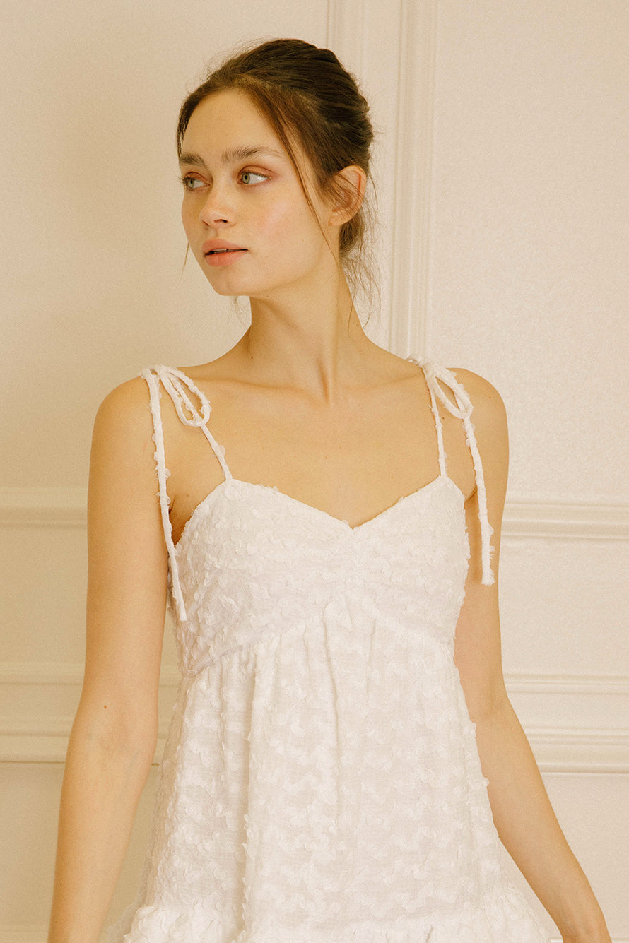 Woman wearing a white dress with adjustable thin straps against a beige wall.