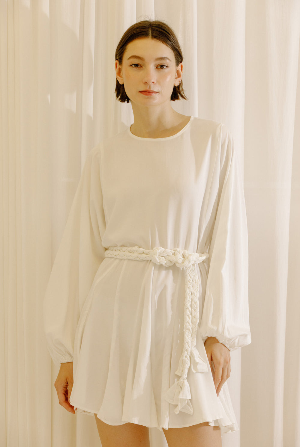 Woman wearing a white dress with long sleeves and a belt against a beige curtain background