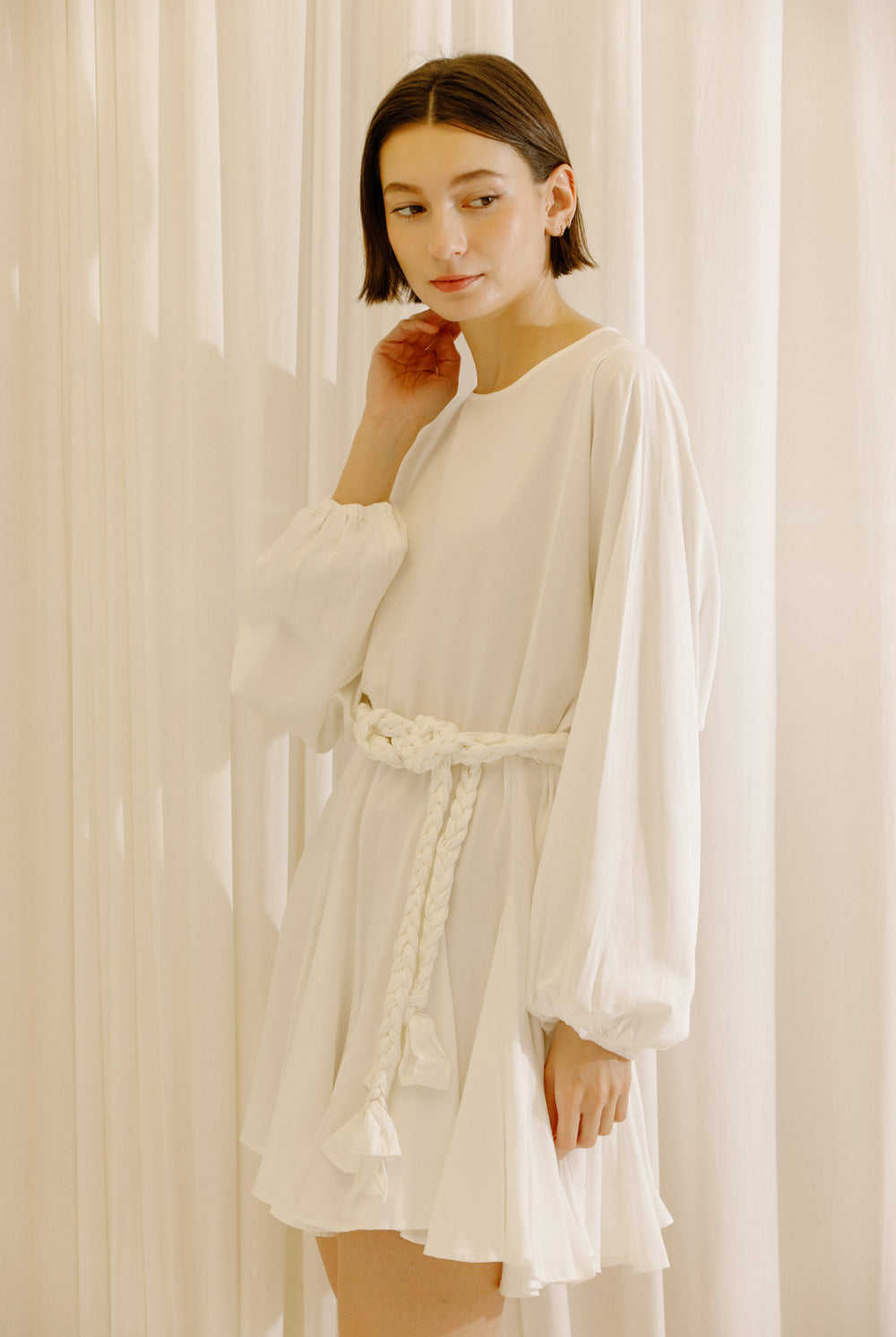 Side view of a woman wearing a white dress with long sleeves and a belt against a beige curtain background