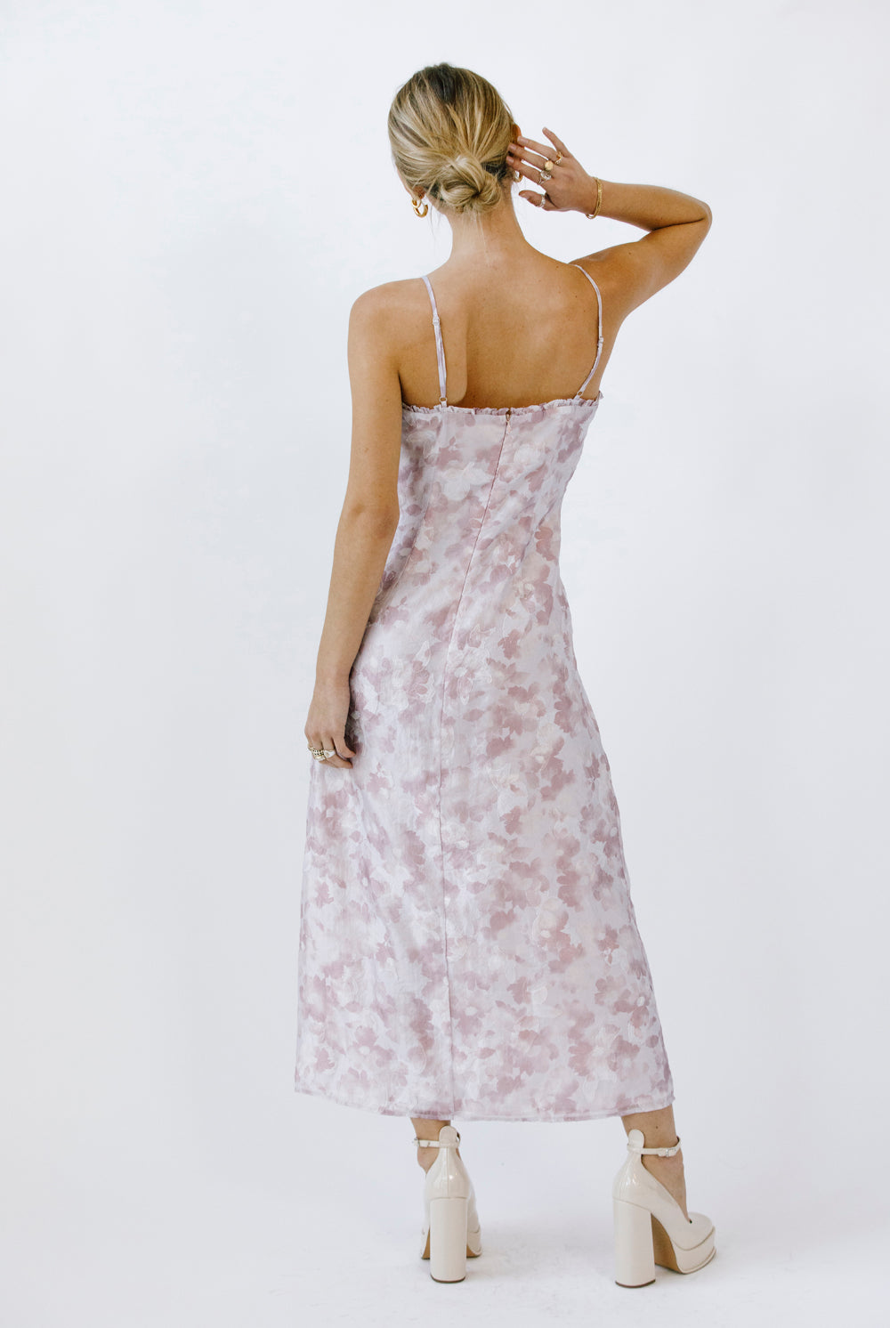Back view of a woman wearing a lilac floral dress with a slit and beige heels on a white background