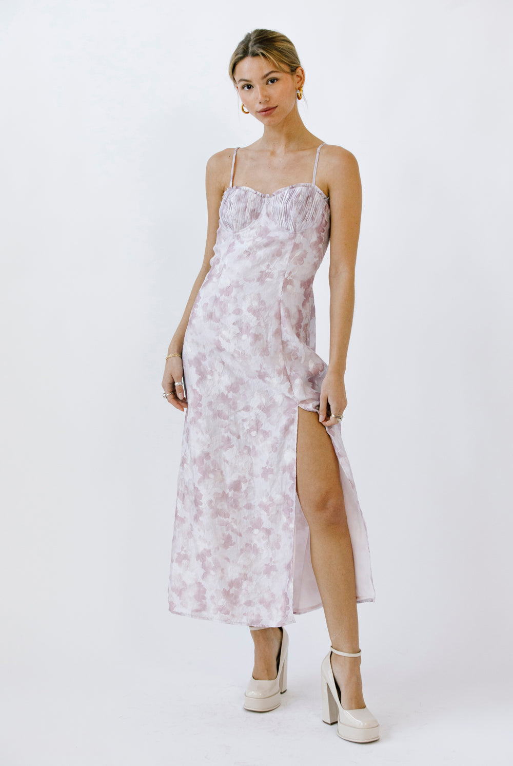 Woman wearing a lilac floral dress with a slit and beige heels on a white background