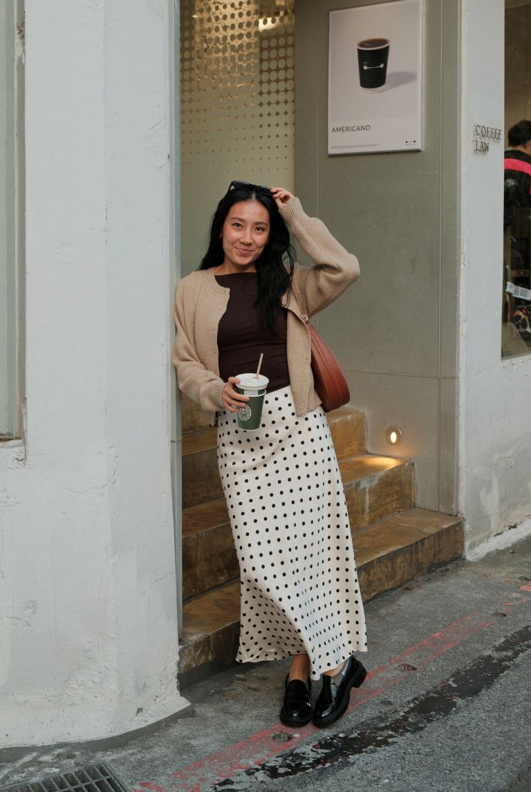 Woman standing outside a building wearing a brown sweater and an ivory and black polka dot skirt, posing for the camera.