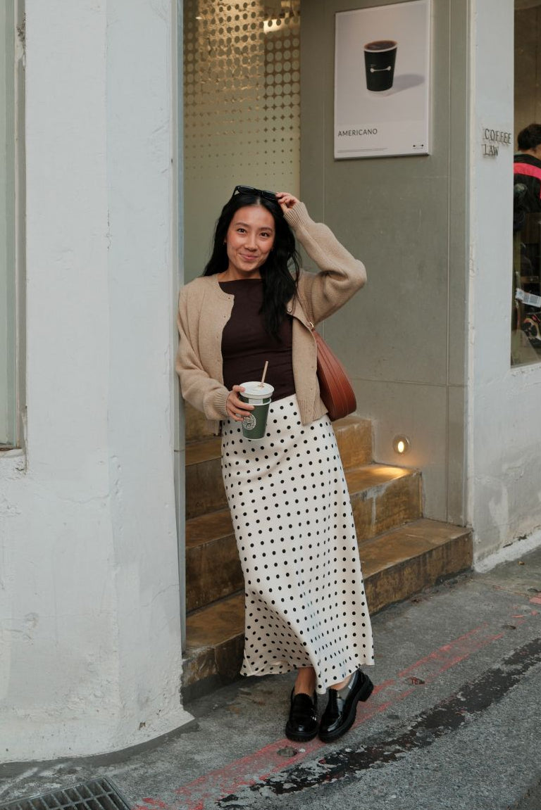 Woman standing outside a building wearing a brown sweater and an ivory and black polka dot skirt, posing for the camera.