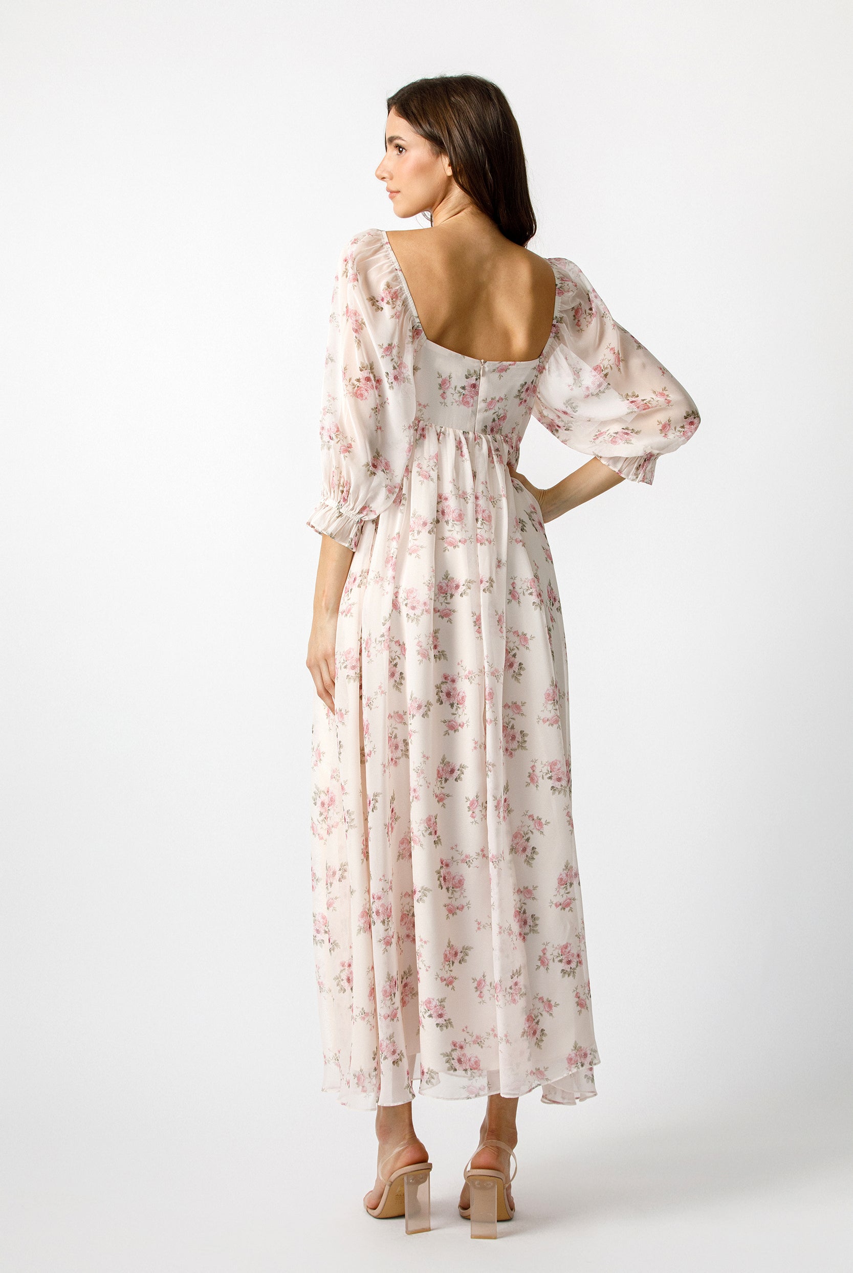 A woman modeling a long-sleeved maxi dress with a pink floral print on a cream background, shown from the back against a light gray studio backdrop.