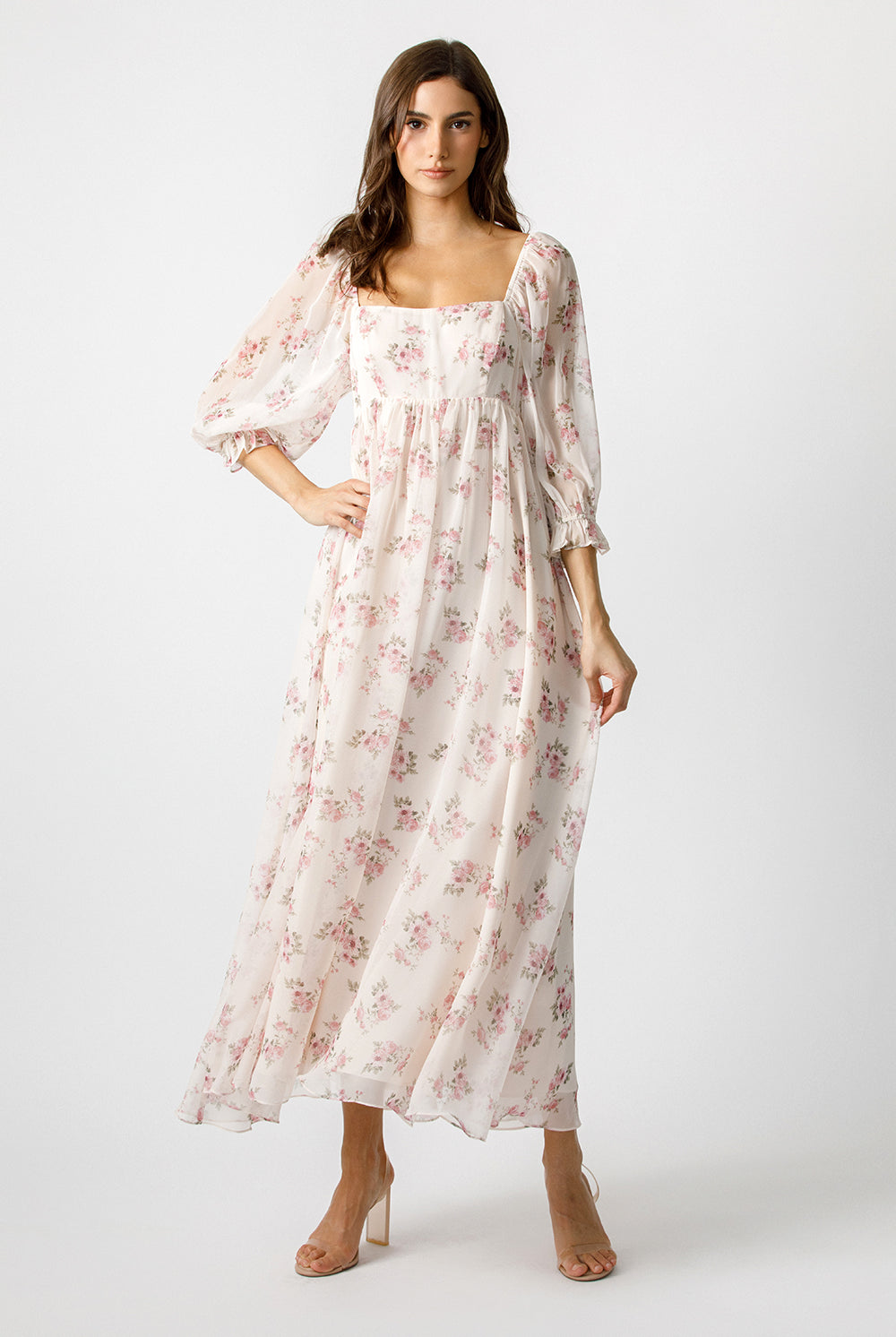 A woman modeling a long, flowy midi dress with a square neckline, puff sleeves, and an allover pink floral print on a light cream background, photographed against a plain light gray studio backdrop.