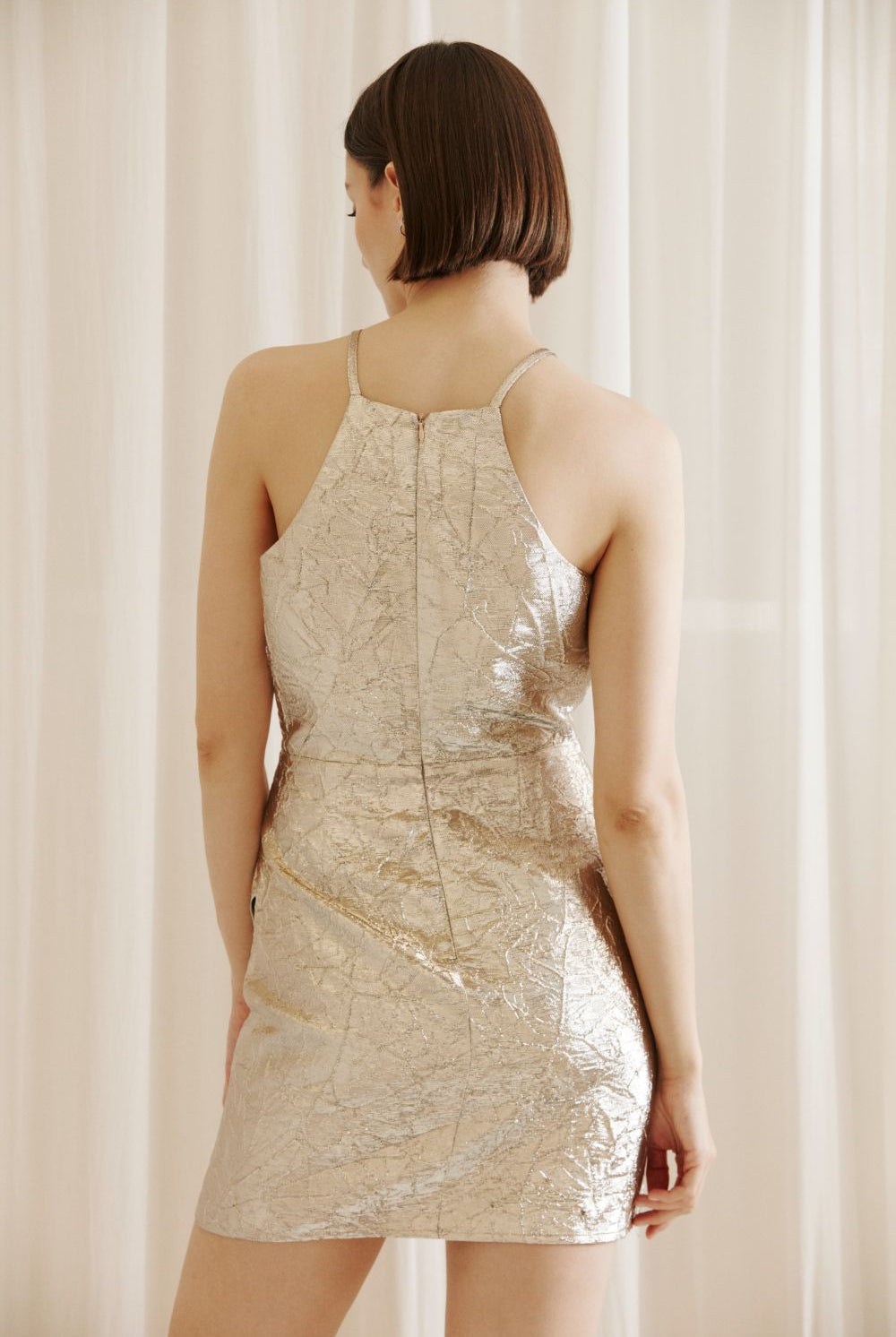 Back of a woman wearing a gold mini dress with an invisible zipper against a beige curtain background