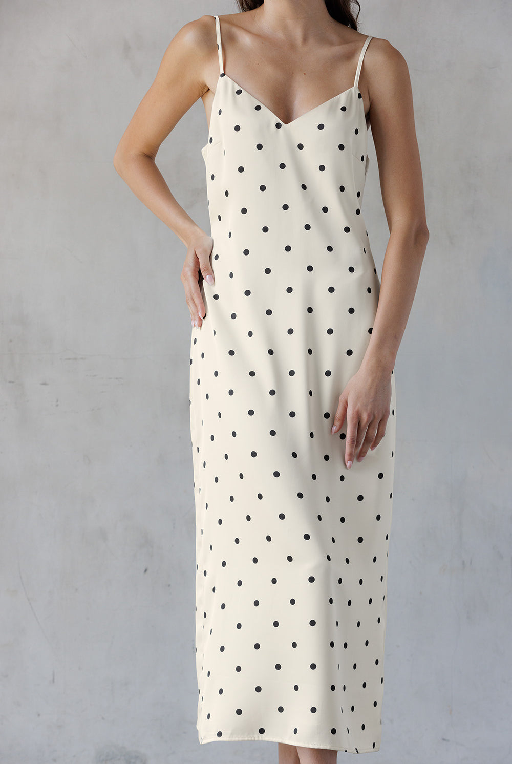 A woman wearing an ivory spaghetti-strap slip dress with a black polka dot pattern, standing against a textured gray wall.