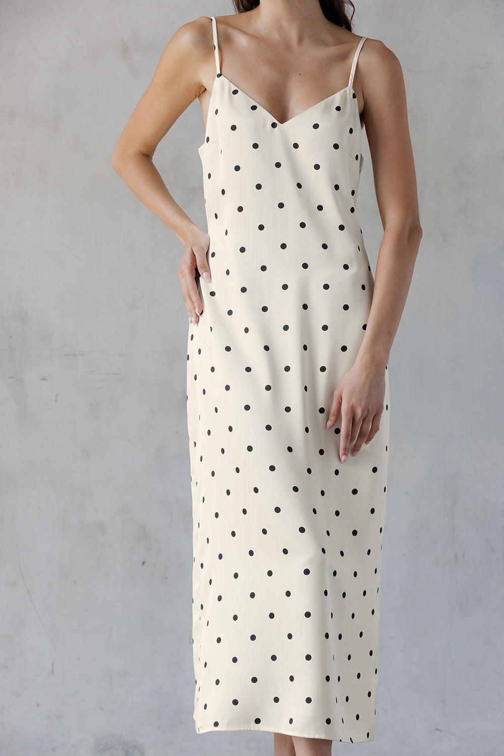 A woman wearing an ivory spaghetti-strap slip dress with a black polka dot pattern, standing against a textured gray wall.