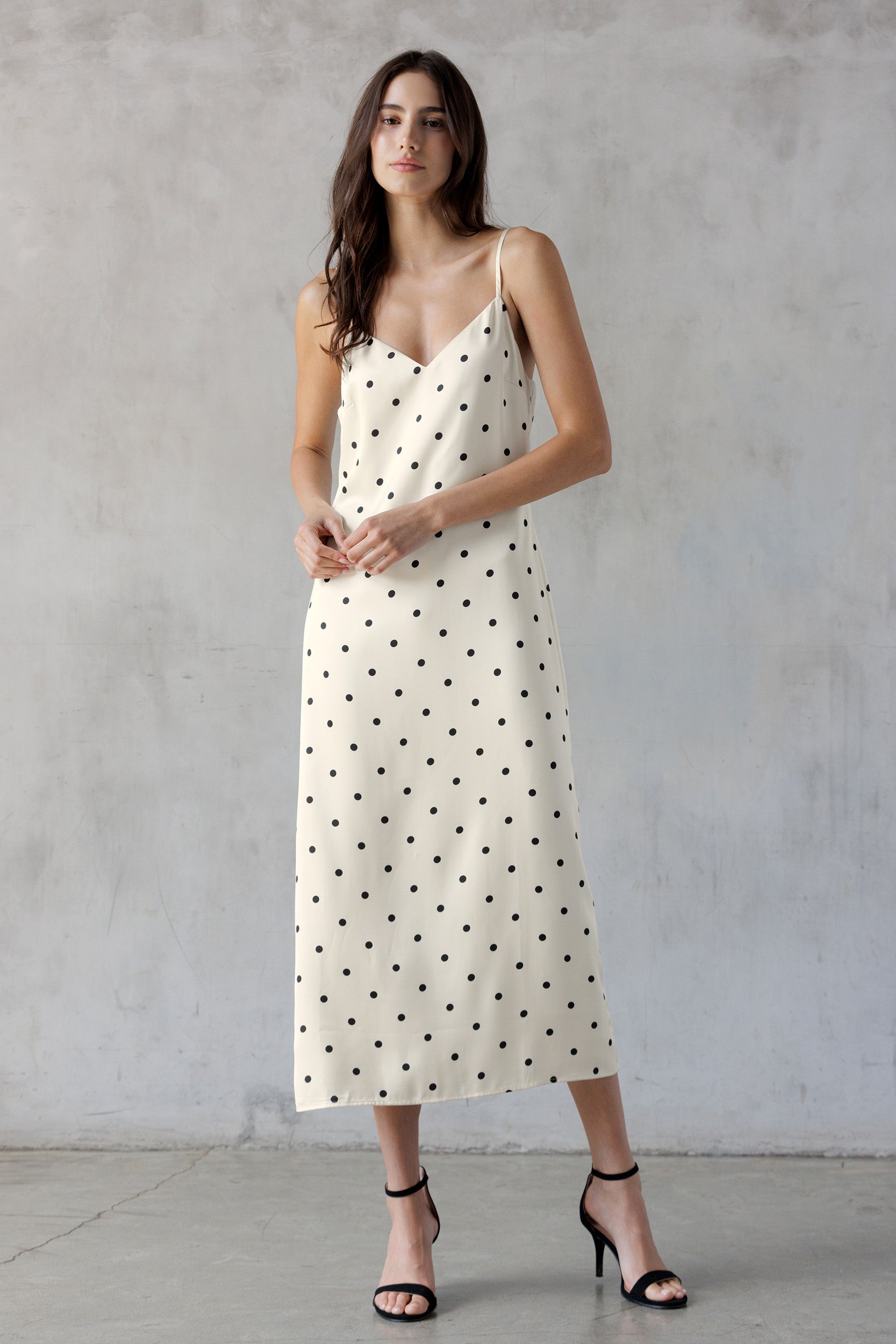 A woman modeling an ivory spaghetti-strap midi dress with black polka dots, paired with black strappy high-heeled sandals, standing against a textured gray concrete wall.