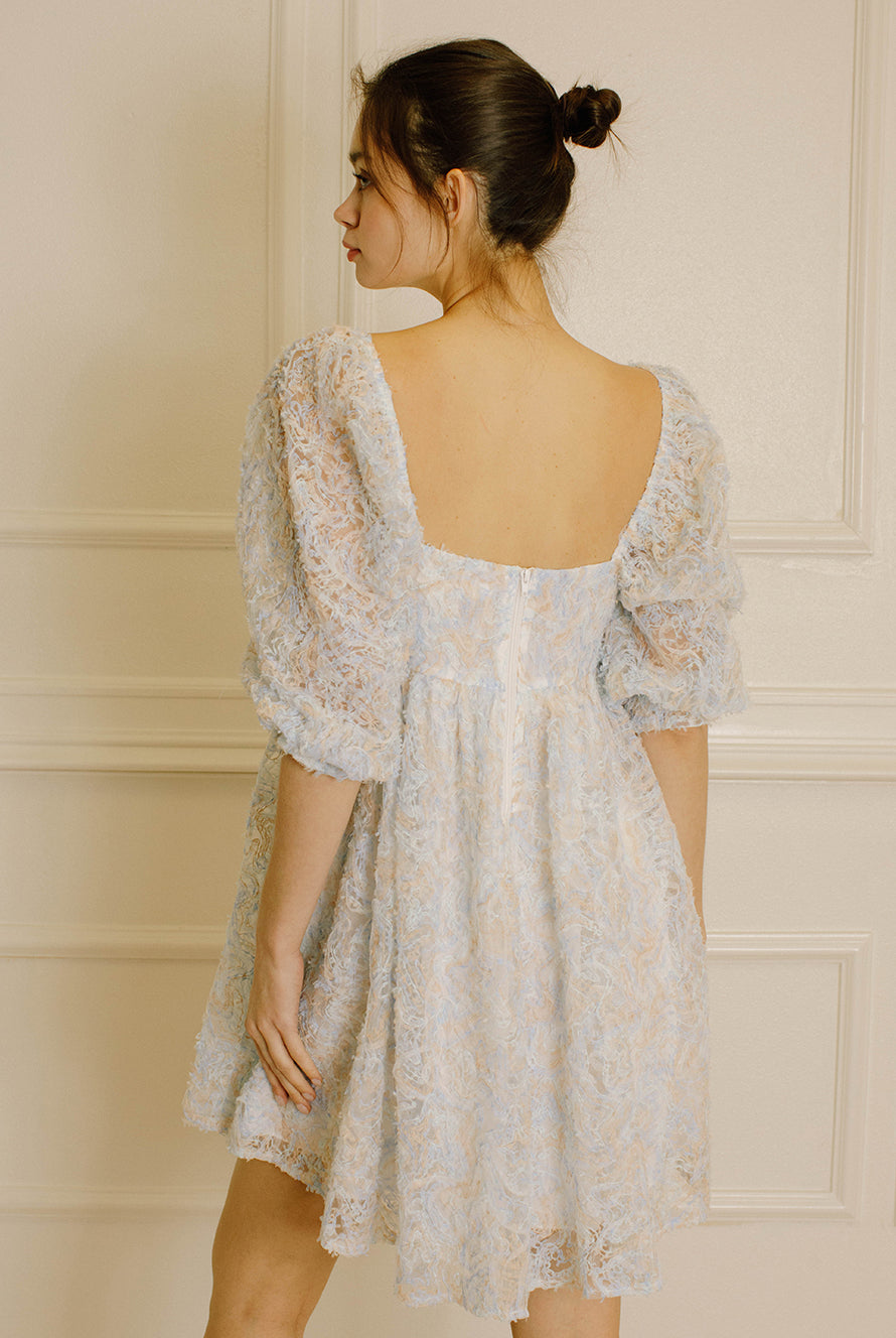 Back view of a woman wearing a blue and cream textured dress against a neutral wall.