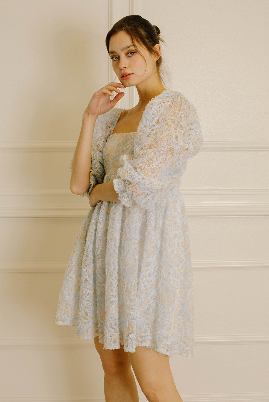 Side view of a woman wearing a blue and cream textured dress against a neutral wall.