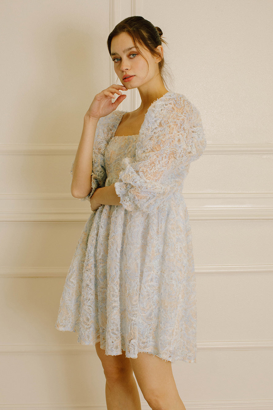 Side view of a woman wearing a blue and cream textured dress against a neutral wall.