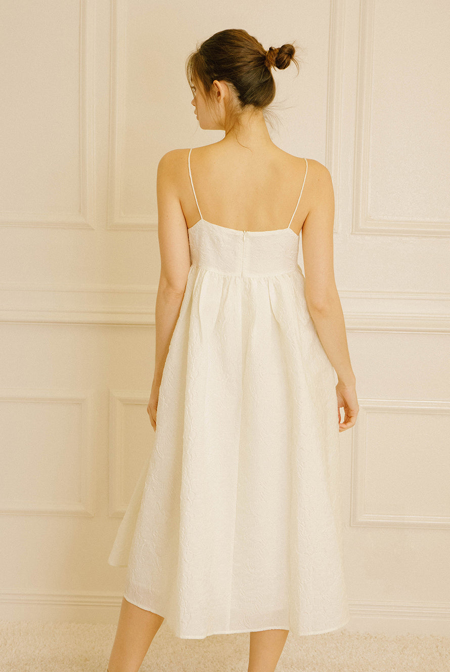 Back view of a woman wearing a cream midi dress with floral jacquard details against a light-colored wall
