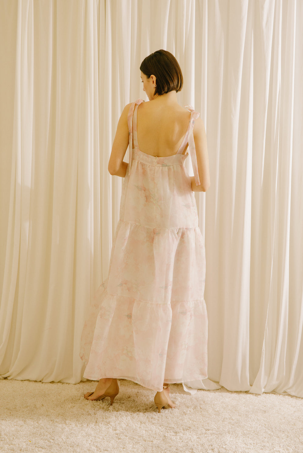 Back view of a woman in a light pink dress standing against a white curtain backdrop